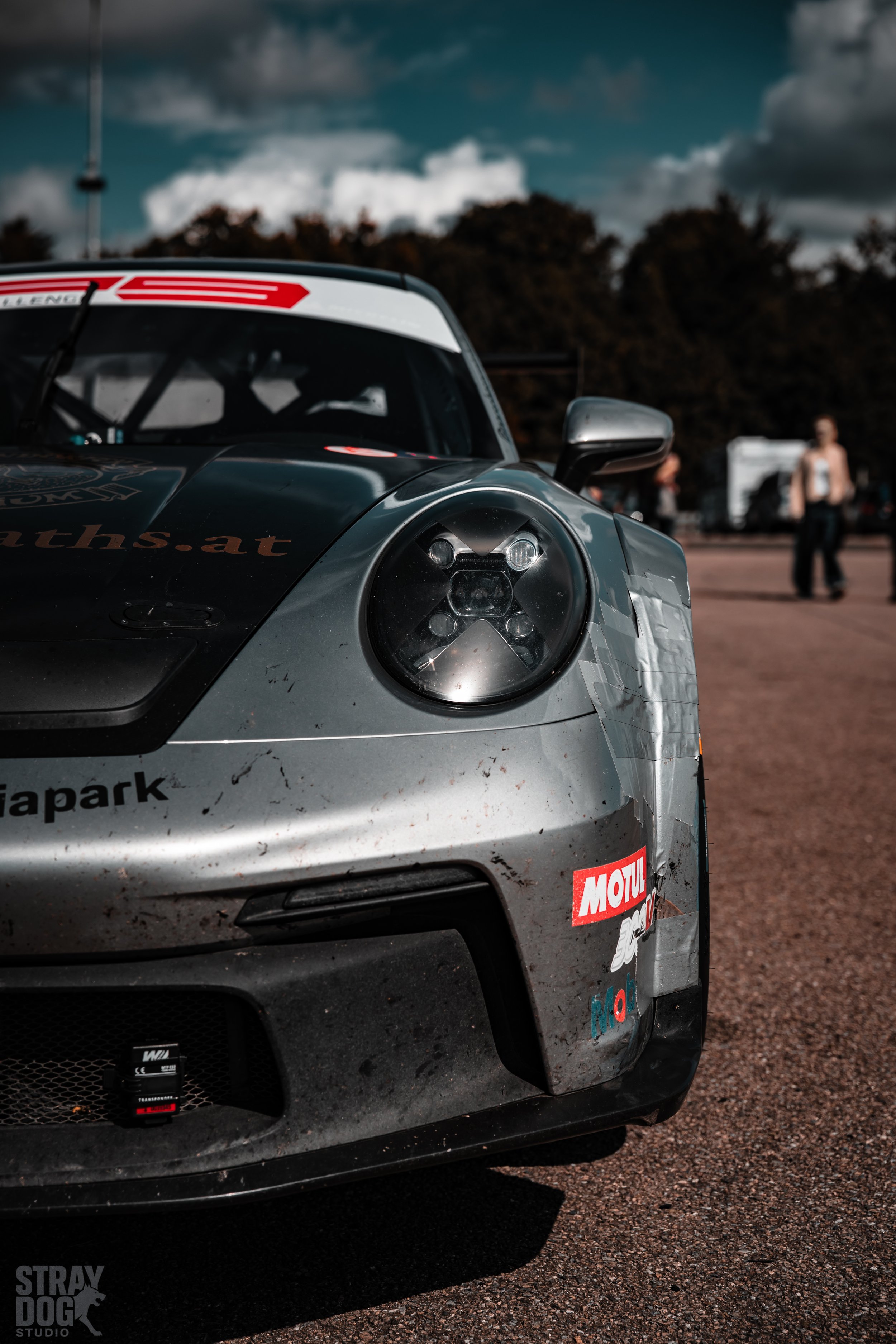 Close-up of a gray racing car with damaged front, dirt, and sponsor stickers, parked on a lot, with trees and people in the background.