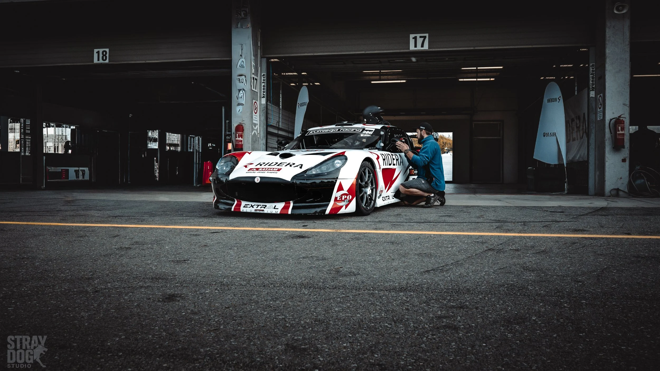 A race car in a pit garage with a person crouching beside it, using a mobile device. The car is white with red and black accents, displaying racing and sponsor logos. The garage has an open shutter, and the ground is paved.