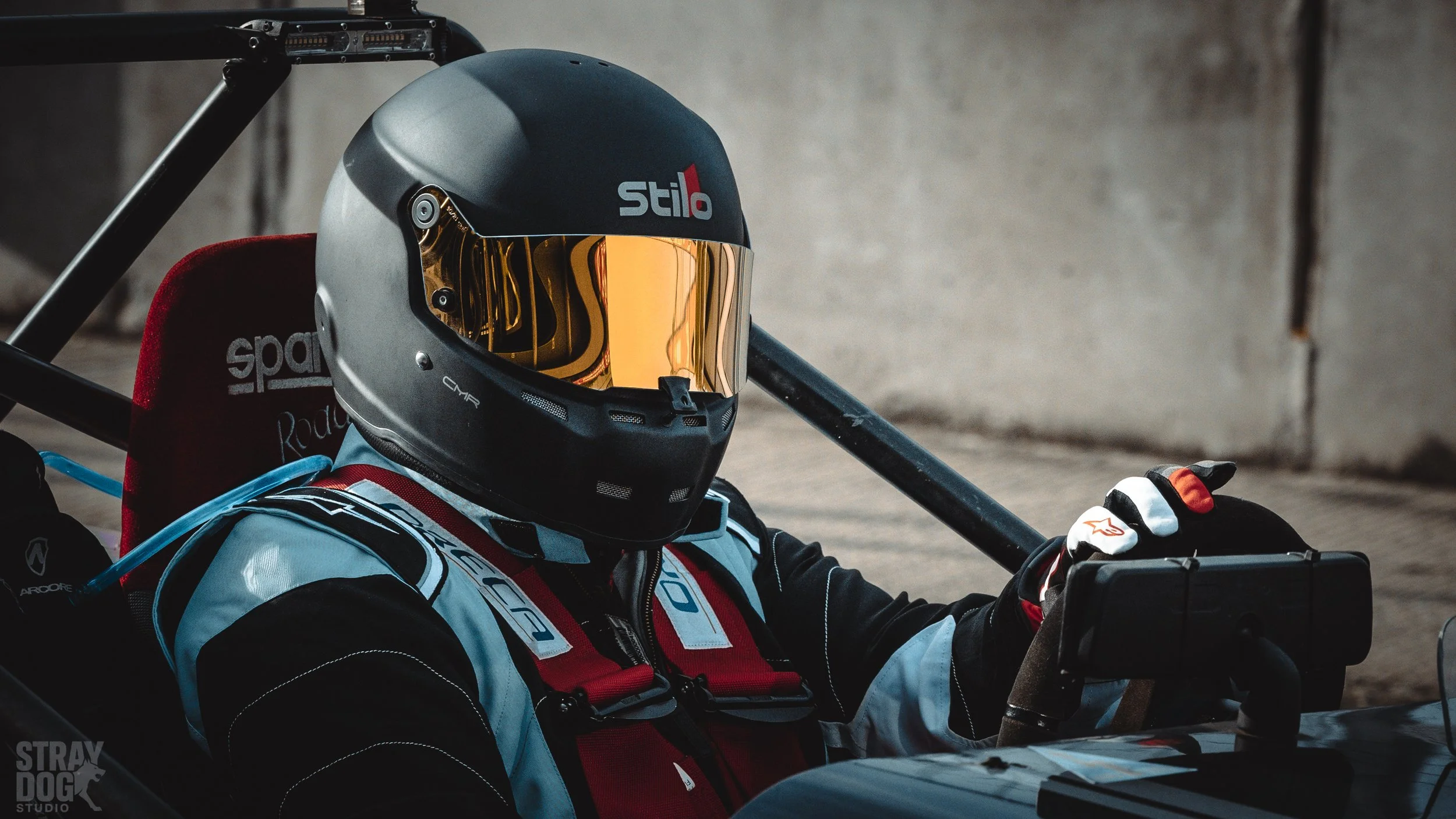 A race car driver wearing a black helmet with a gold tinted visor, a padded red and black racing suit, and gloves, sitting inside a race car with a black roll cage and a red headrest.