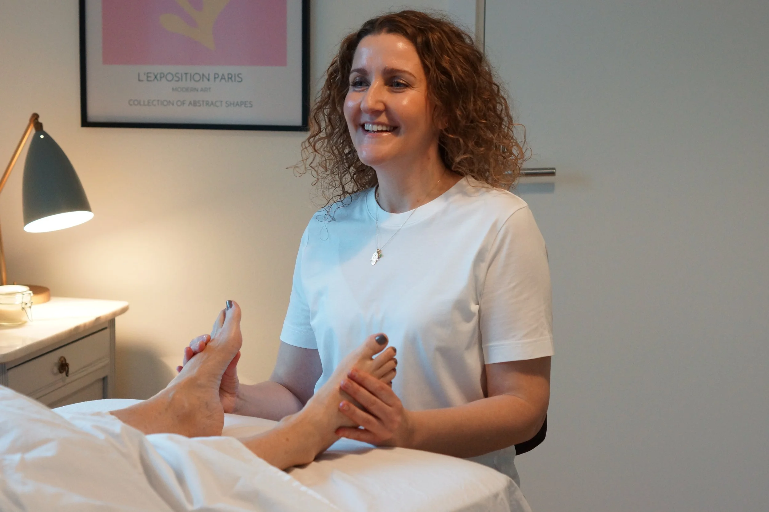 A woman with curly red hair smiling and holding hands with a person lying in bed. The person in bed has a white blanket and is reaching out to hold her hands. There is a lamp and a framed art piece on the wall in the background.