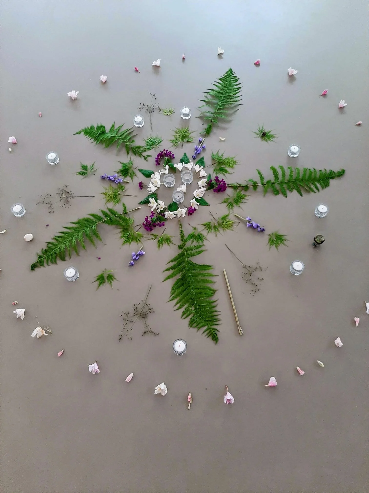 A flat surface decorated with green fern leaves, purple and white flowers, small candles, and scattered pink flower petals.
