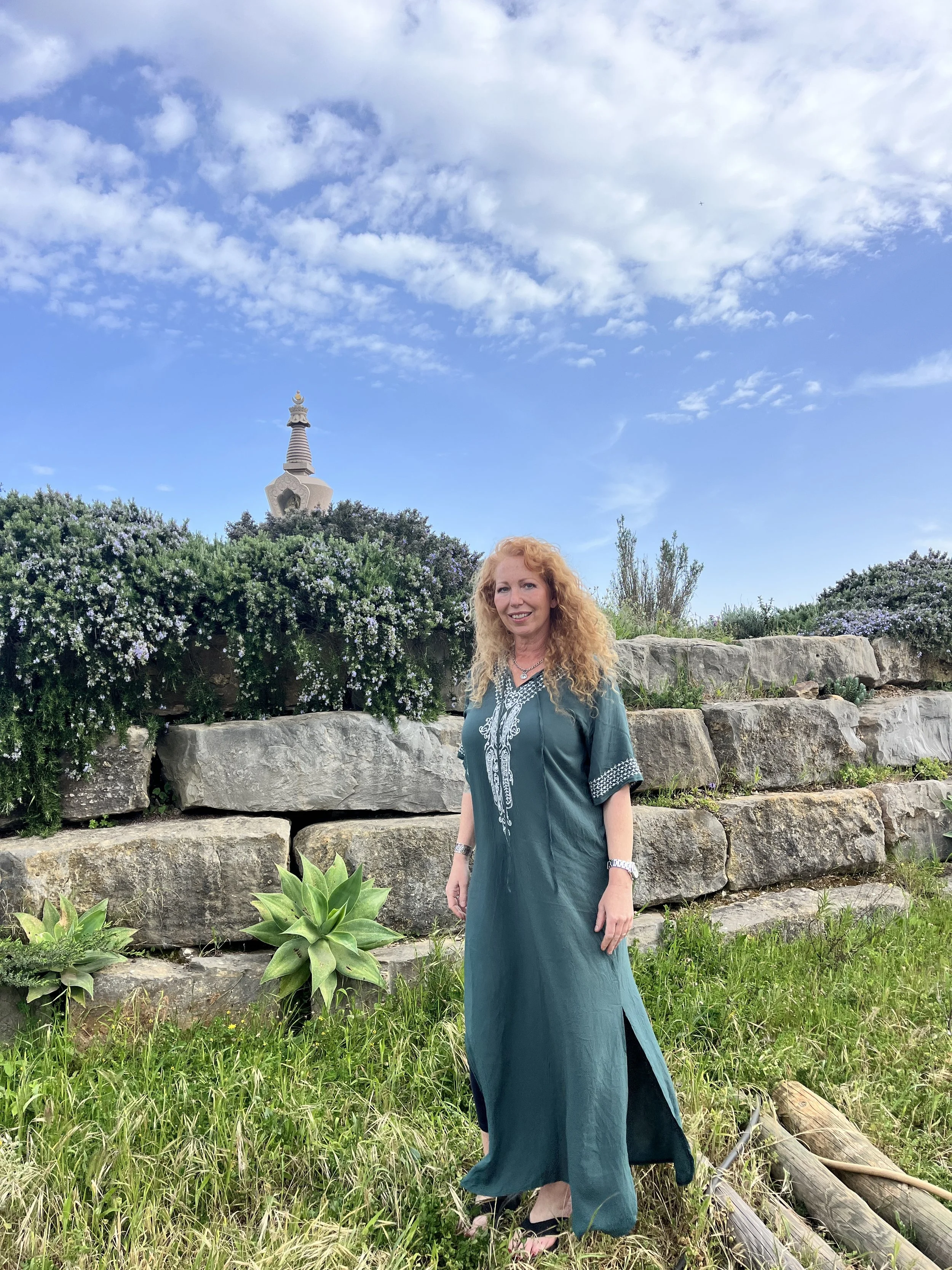 A woman with curly red hair wearing a teal traditional dress with embroidered white designs, standing outdoors in a garden with green plants, large rocks, and bushes with white and purple flowers. In the background, there is a Buddhist stupa with a golden spire, under a bright blue sky with scattered clouds.