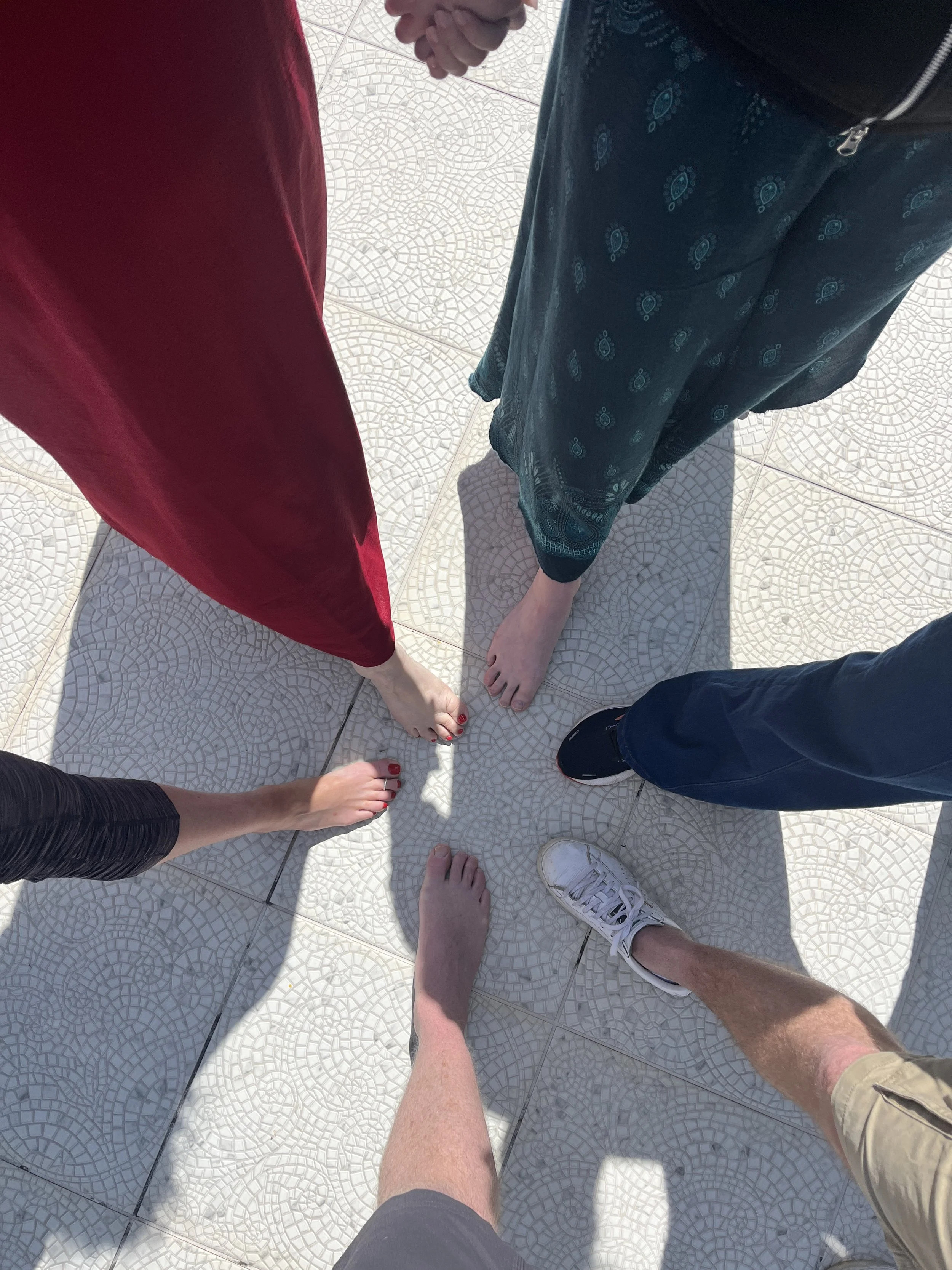 Six people standing on a tiled floor, holding hands in a circle. The image is taken from above, showing their bare feet and part of their legs, with the hands in the center.