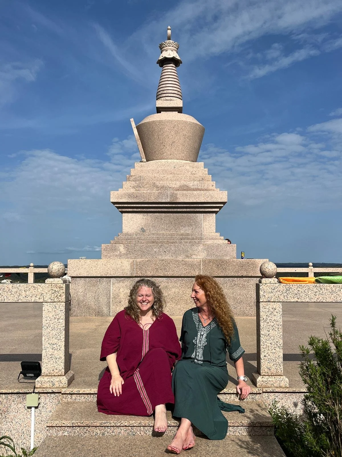 Two women sitting on a stone bench in front of a large monument or sculpture resembling a chess piece or spire, with a blue sky and scattered clouds in the background.