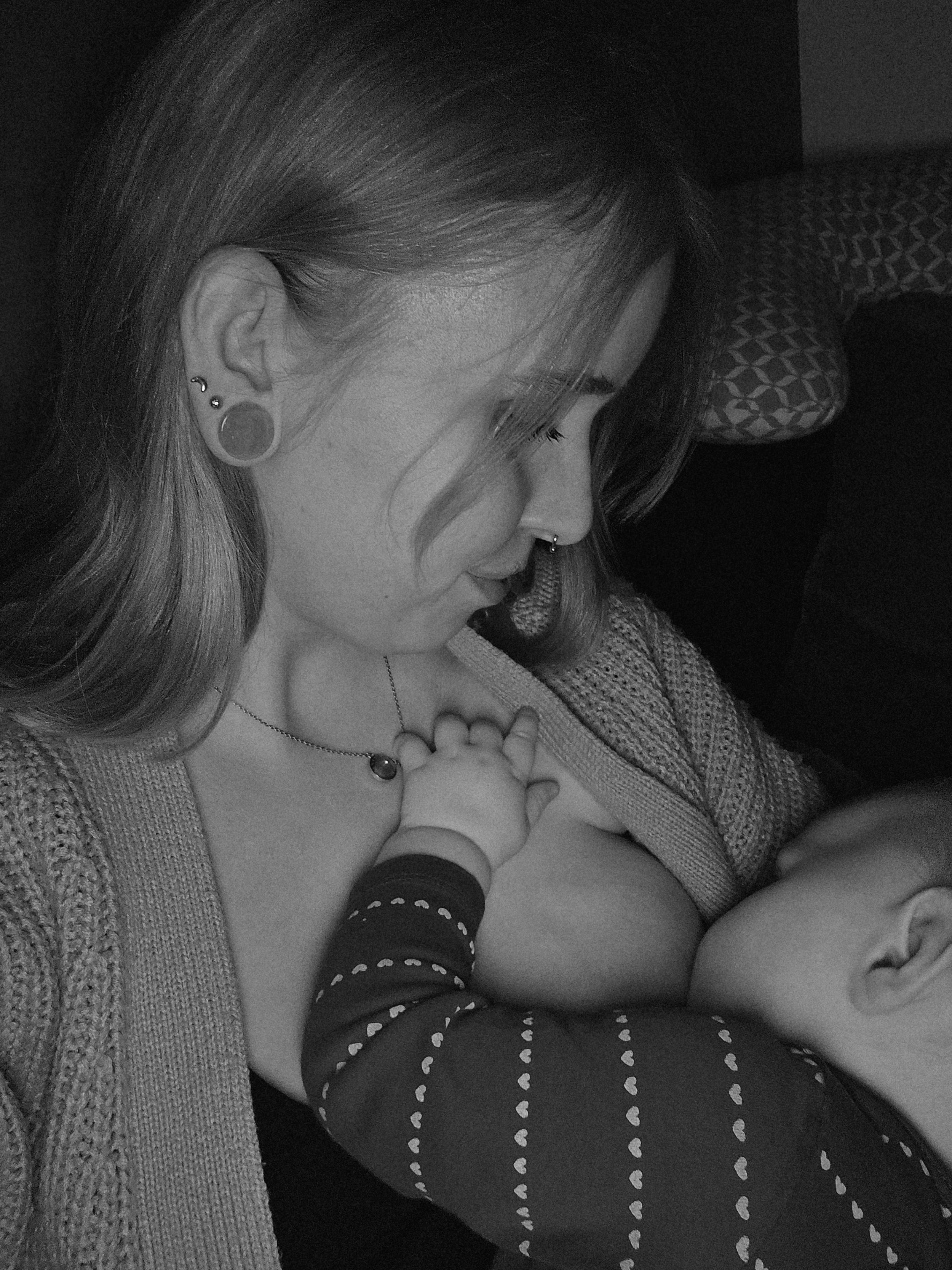 Eine Frau, die ein Baby stillt, in einer schwarzen und weißen Fotografie. Die Frau trägt Schmuck und hat lange Haare.