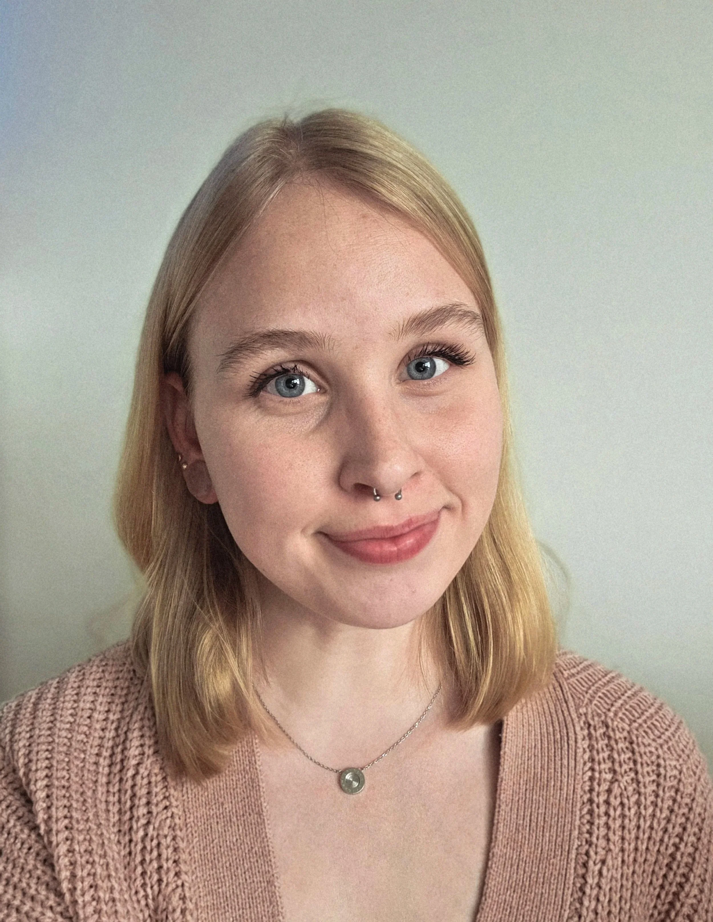 Porträt einer jungen Frau mit blonden, schulterlangen Haaren und blauen Augen, trägt einen rosa Pullover und eine silberne Halskette, vor einem hellen Hintergrund.