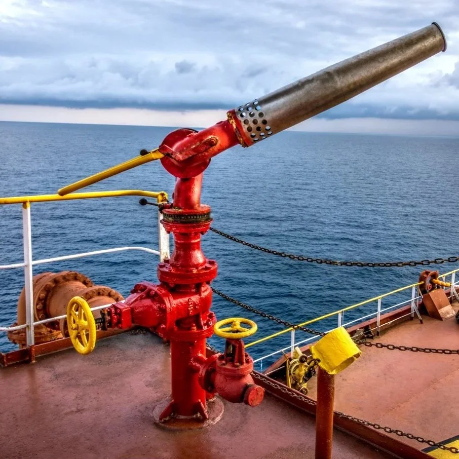 View of a ship's deck with a large brass fire monitor aimed over the ocean, yellow and red valves, chains, and safety features.