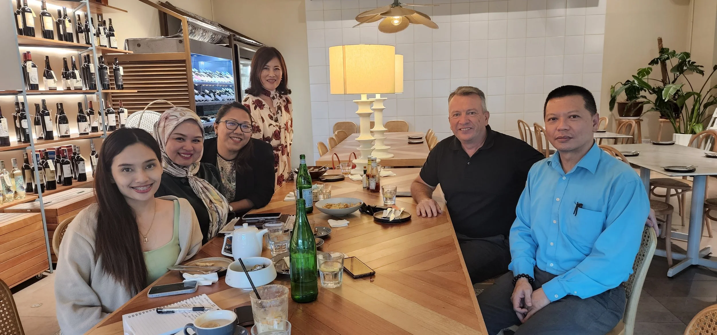 A group of six people, three women and three men, sitting at a restaurant table with food and drinks, smiling for the camera. The restaurant has a modern interior with wine bottles on shelves and potted plants.