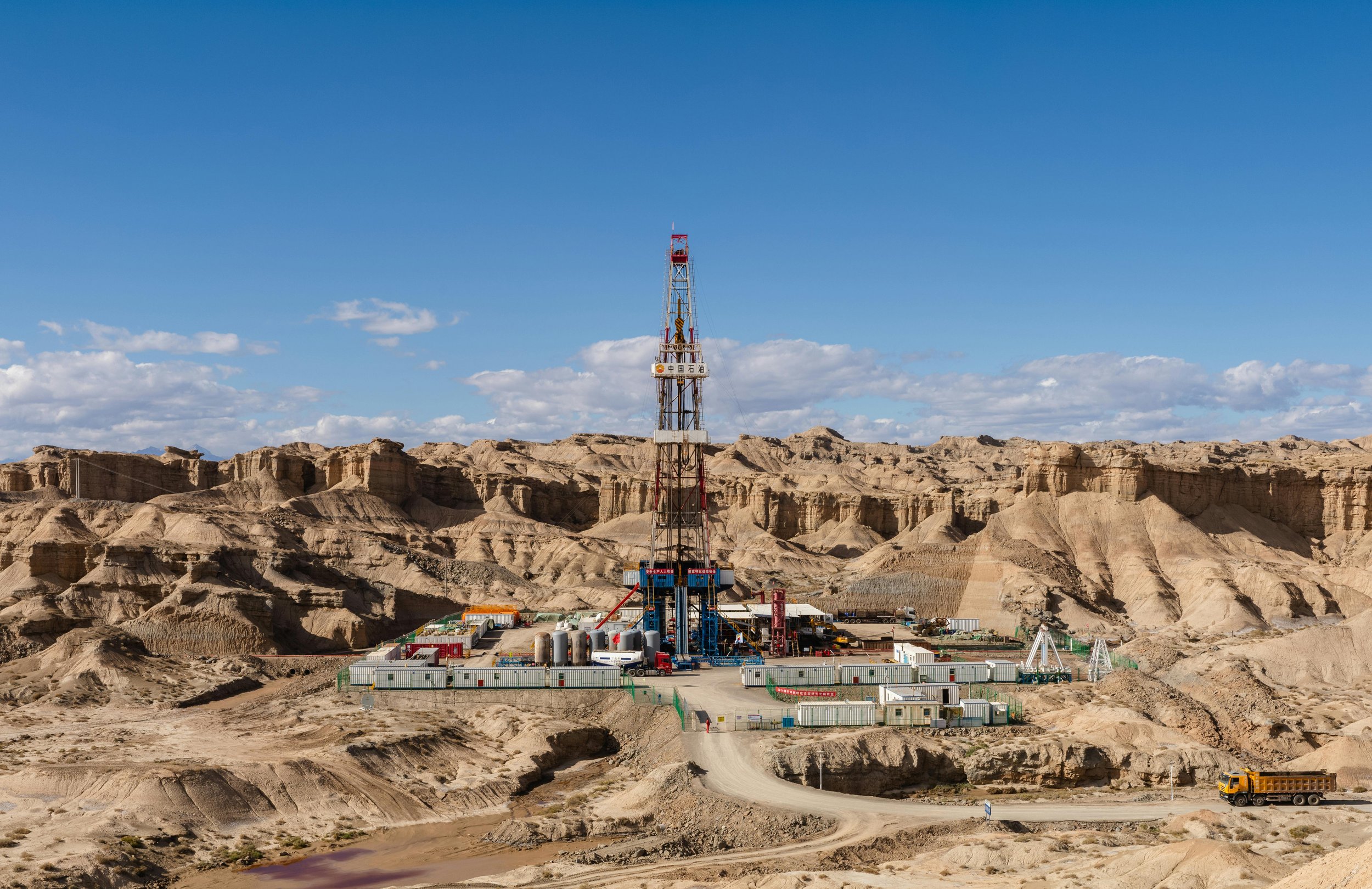 Oil drilling rig in a desert landscape with rugged hills and a blue sky.