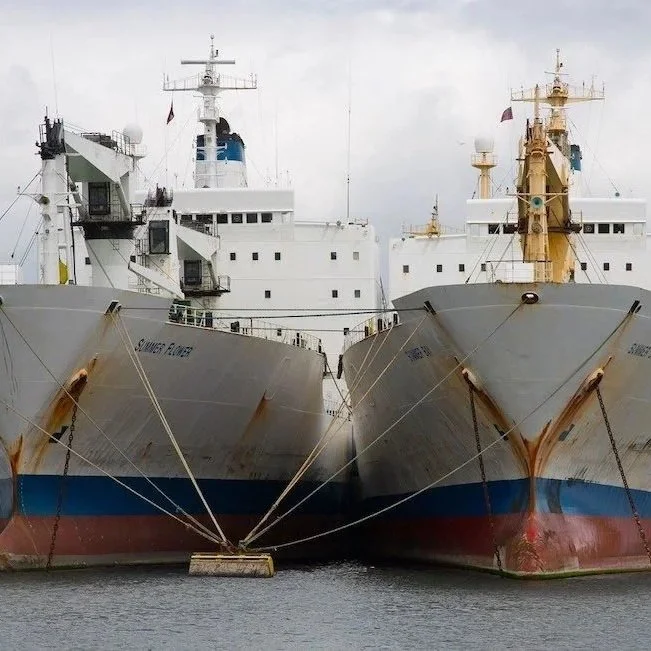 Two large ships docked side by side in a harbor, with rust marks visible on their hulls.