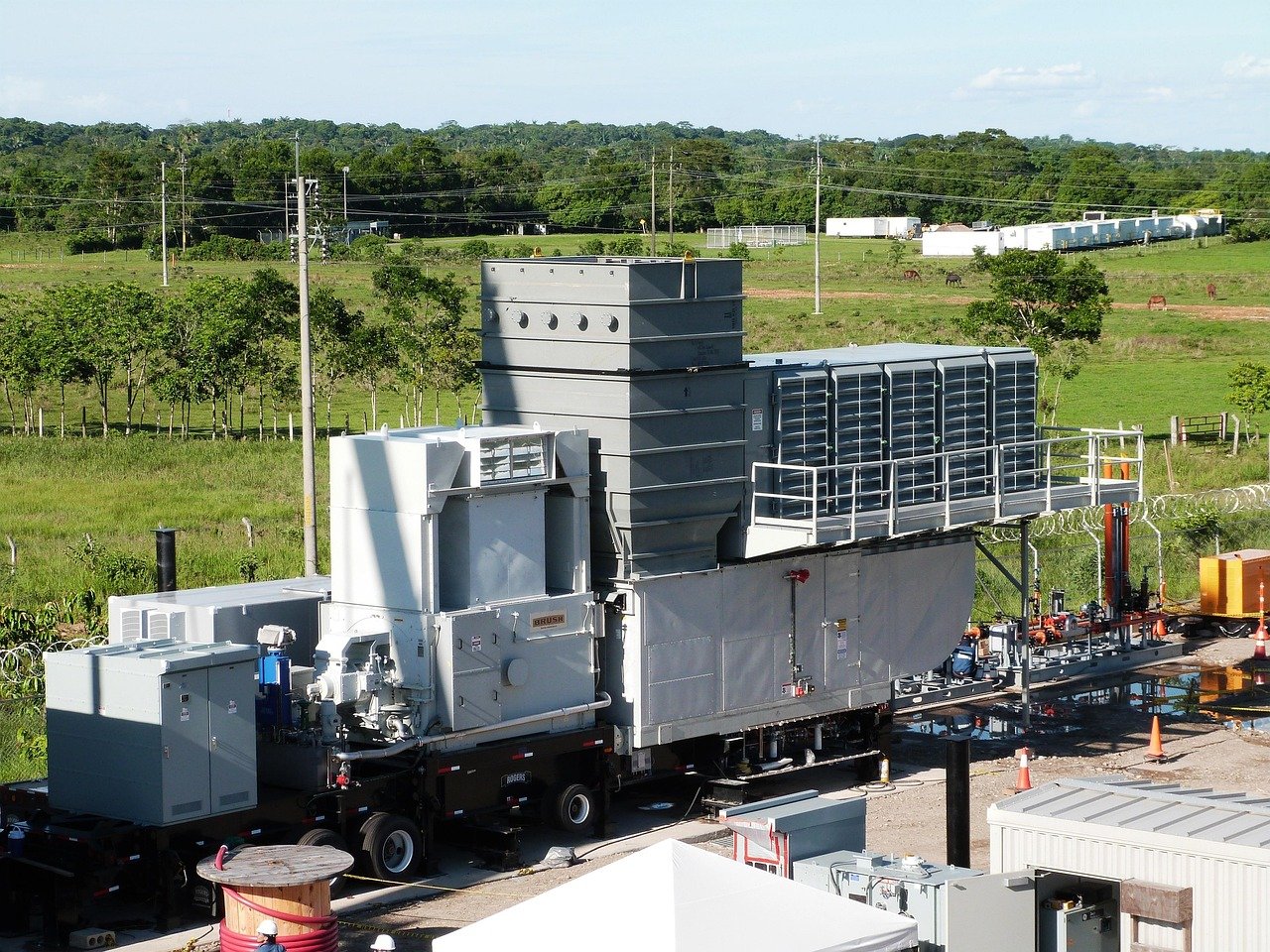 A large industrial machine or generator on a trailer with surrounding equipment, set outdoors in a rural area with fields, trees, and utility poles in the background.