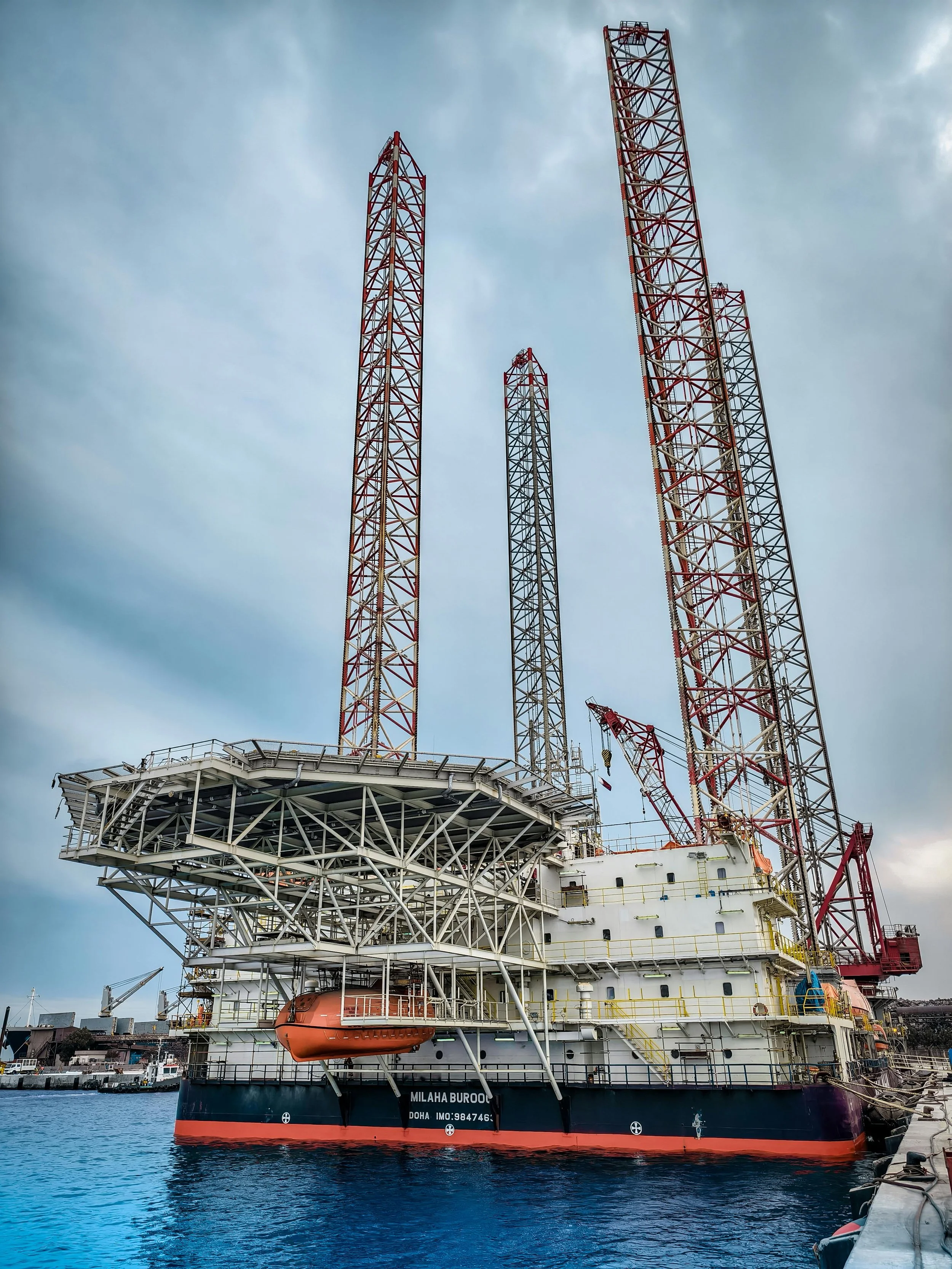Offshore oil drilling rig in harbor with multiple tall red and white towers and a blue sky overhead.