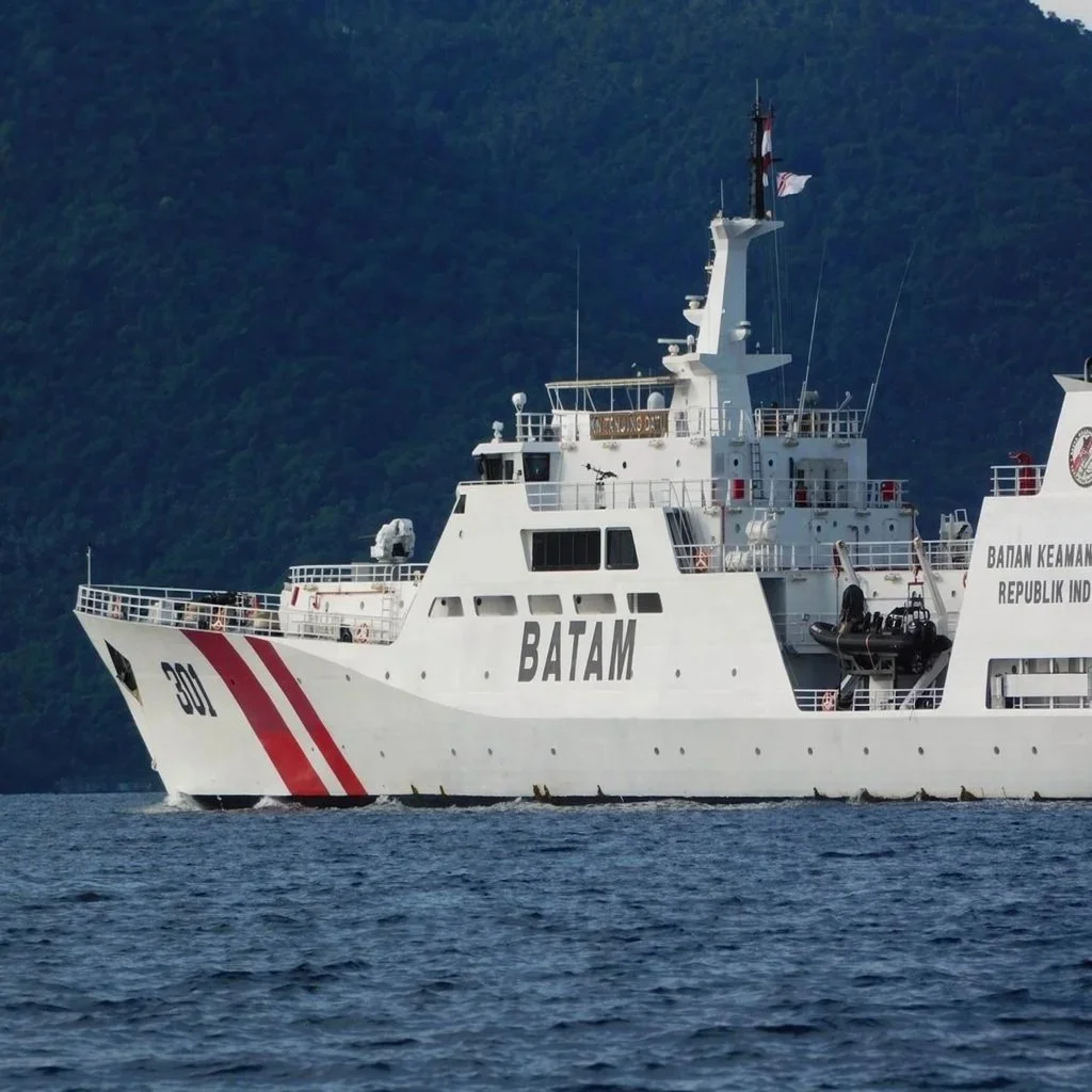 A white naval ship with red and black markings, labeled 'BATAM,' sailing on the water with a backdrop of green mountains.