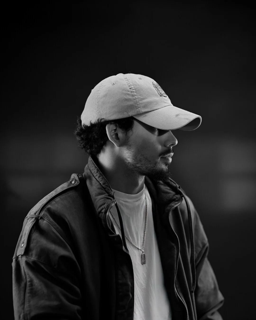 Black and white profile of a young man wearing a baseball cap, jacket, and necklace, looking downward.