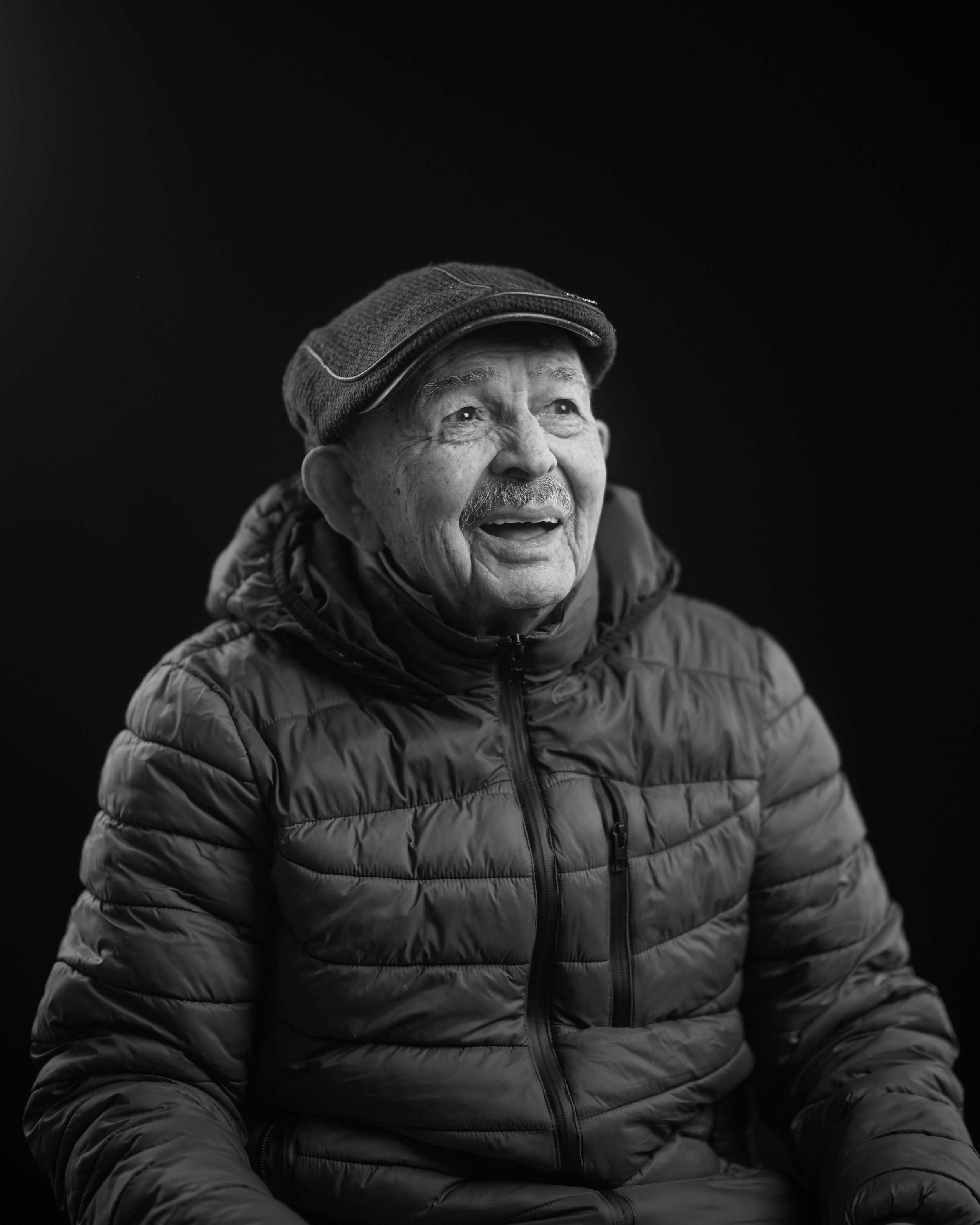Black and white photo of an elderly man smiling, wearing a patterned cap, a puffy jacket, and sitting against a dark background.