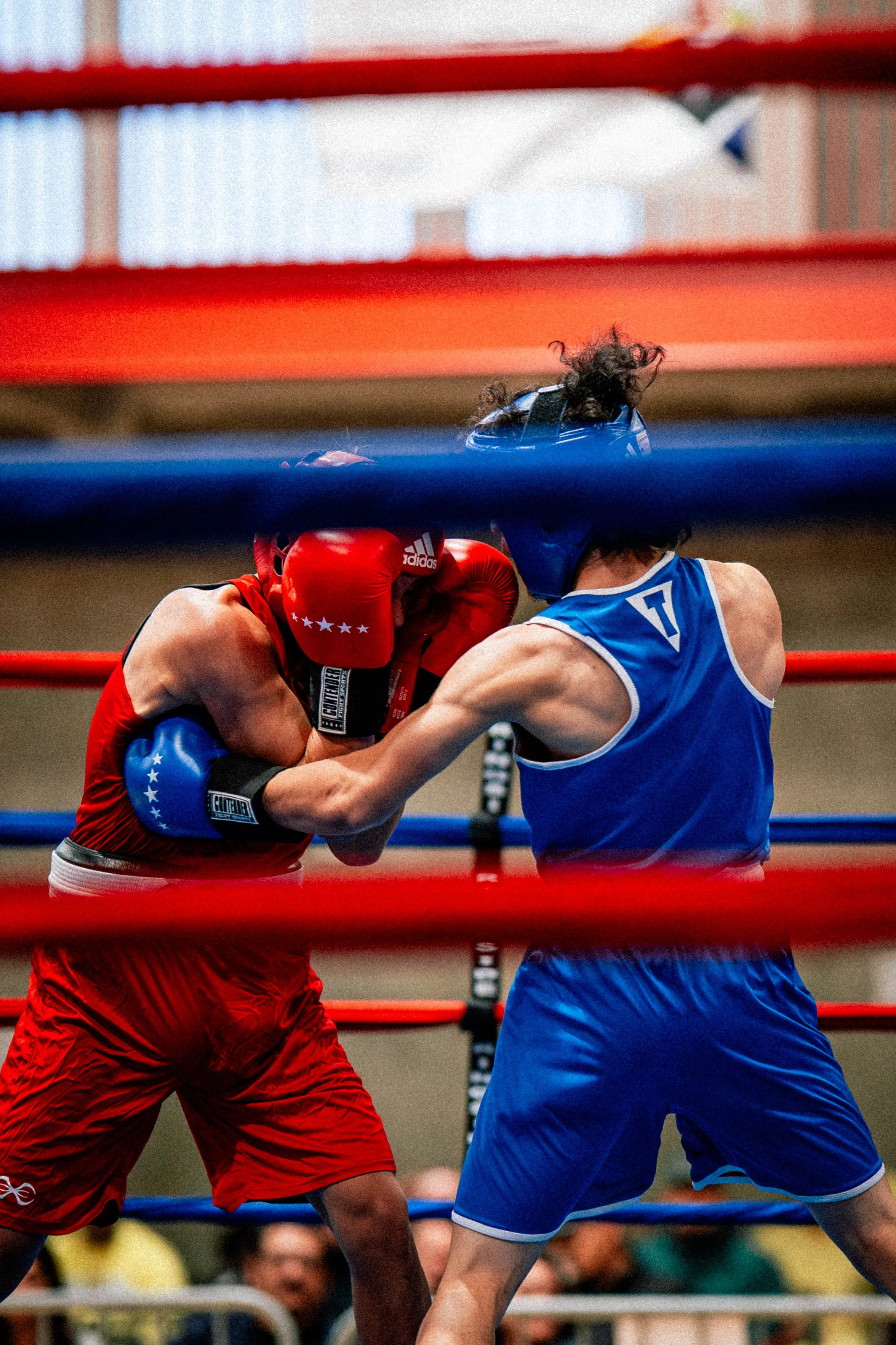 Two amateur boxers in red and blue protective gear sparring in a boxing ring, with red and blue ropes visible.