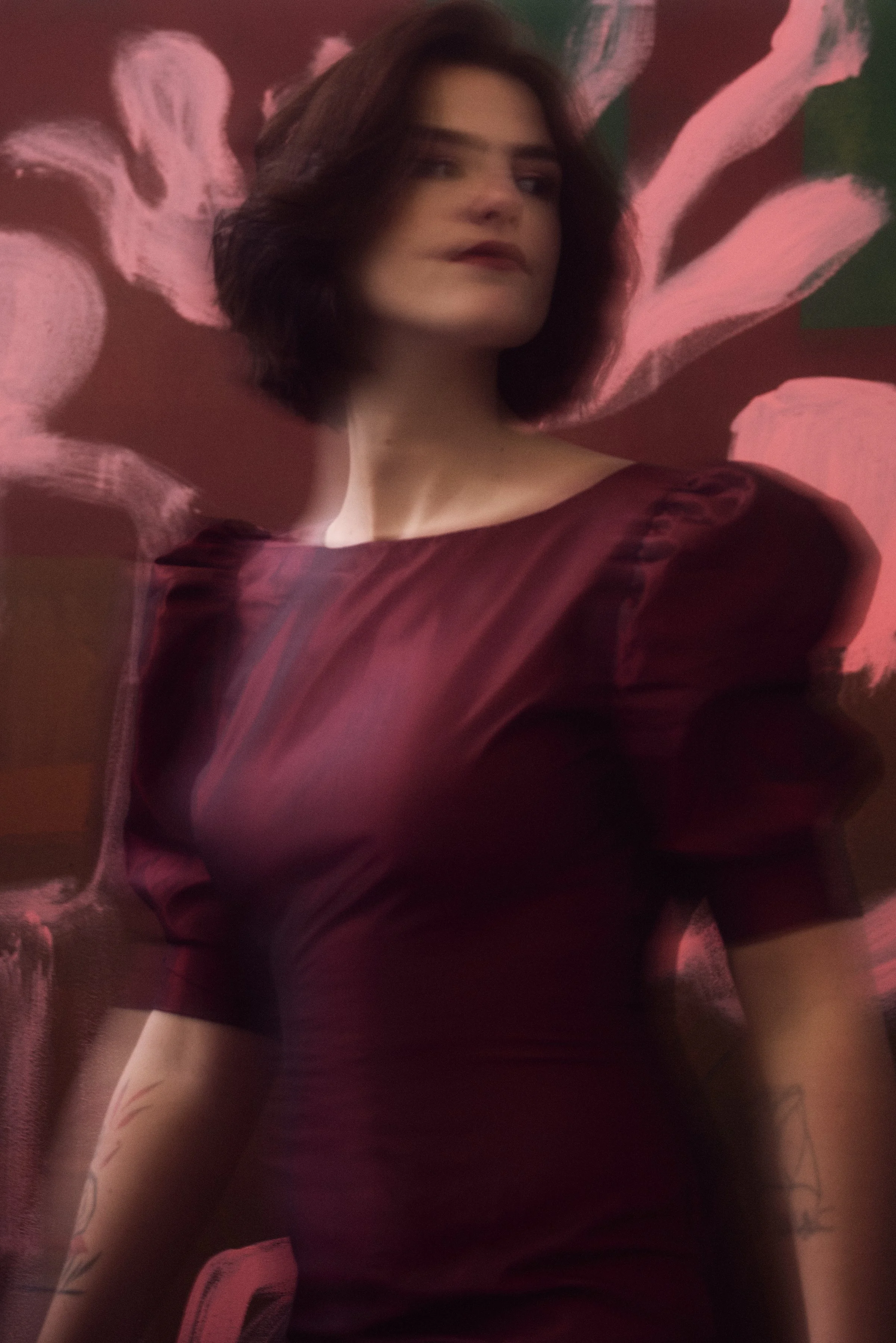 A woman with shoulder-length dark hair wearing a dark red satin dress with puffed sleeves, standing against a pink and green floral painting or backdrop.