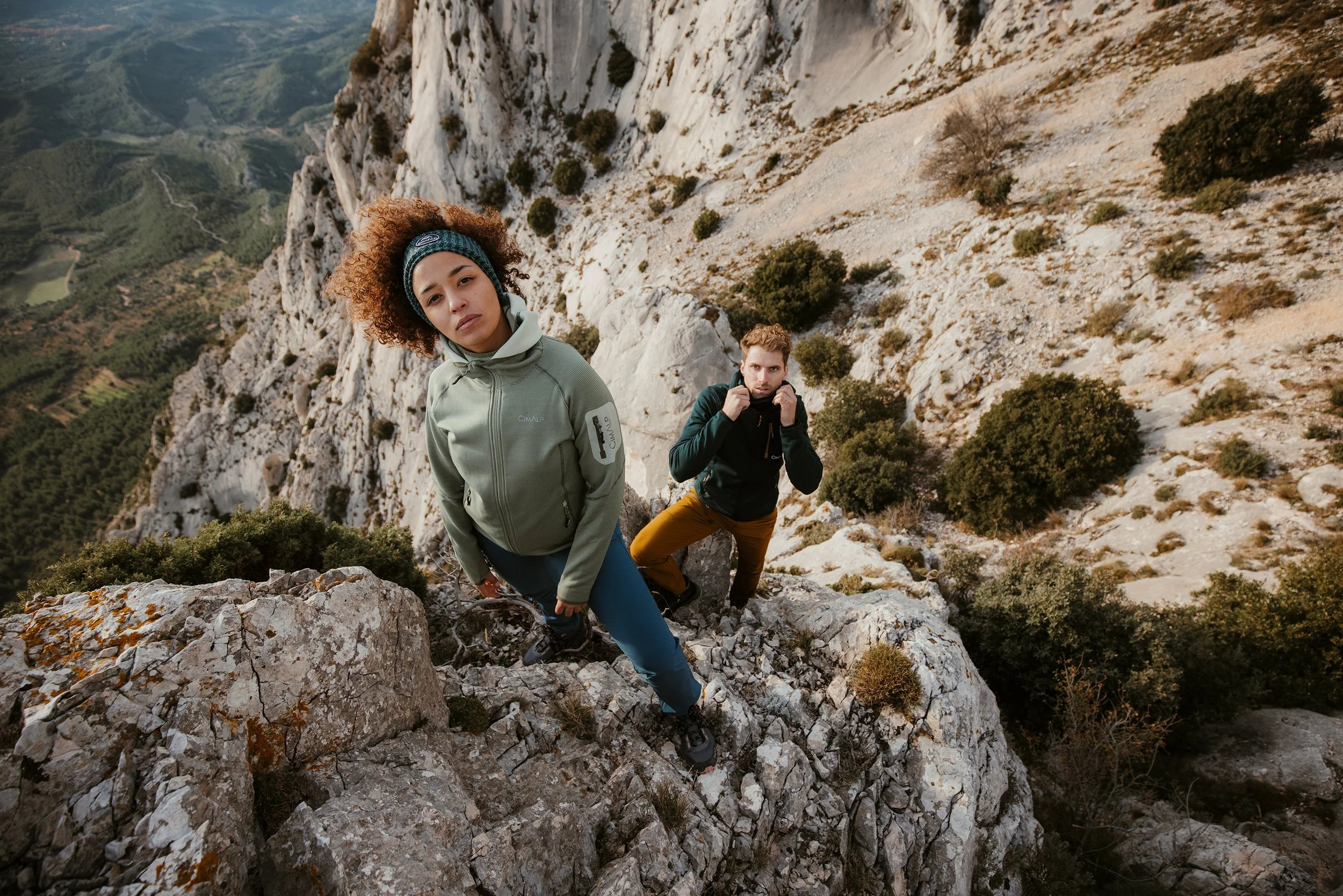 Deux personnes escaladant une falaise rocheuse dans un paysage montagneux avec des buissons et des montagnes en arrière-plan.