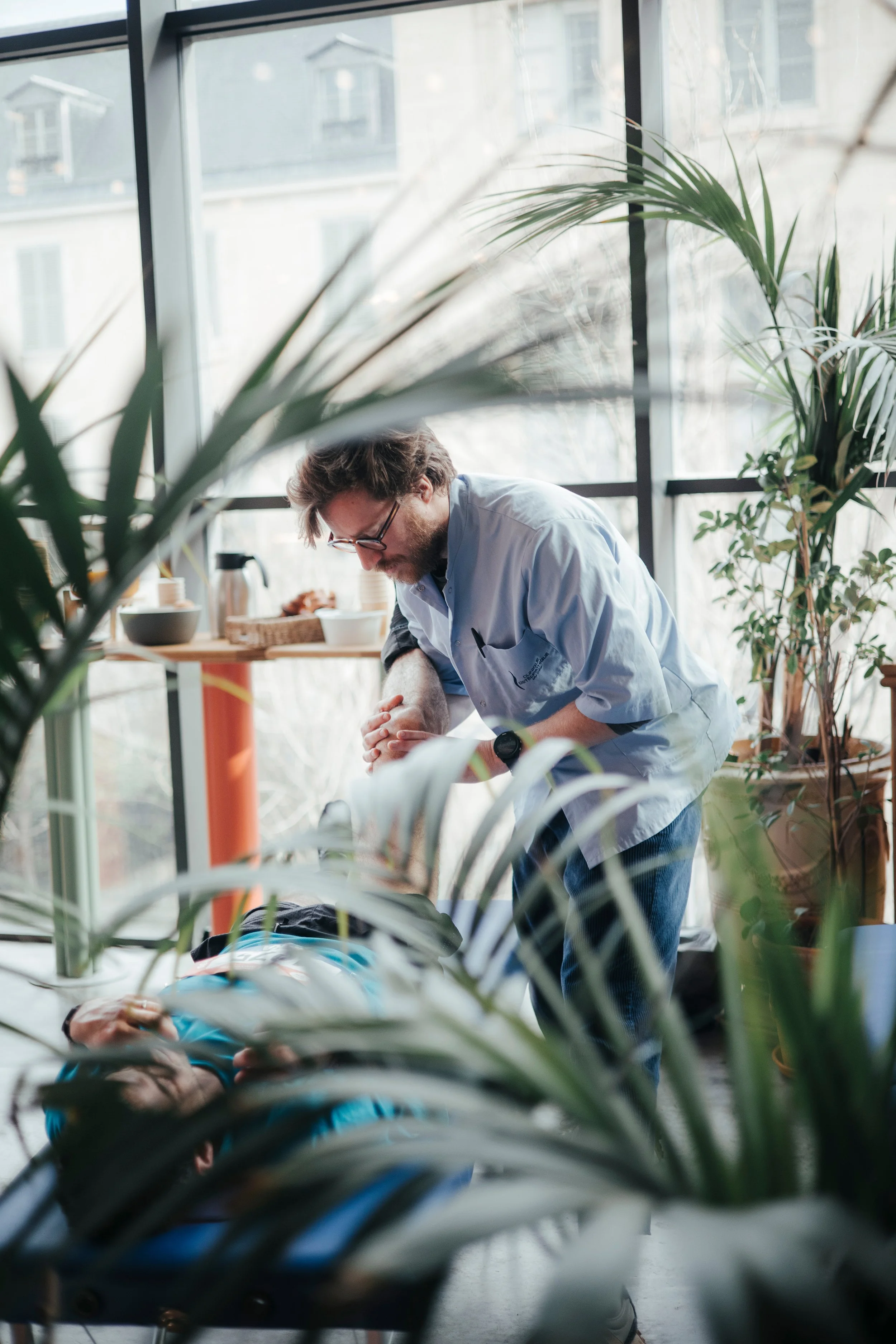 Un secouriste pratique le massage cardiaque sur une victime allongée sur un lit de soins, dans un endroit entouré de plantes et de fenêtres lumineuses.