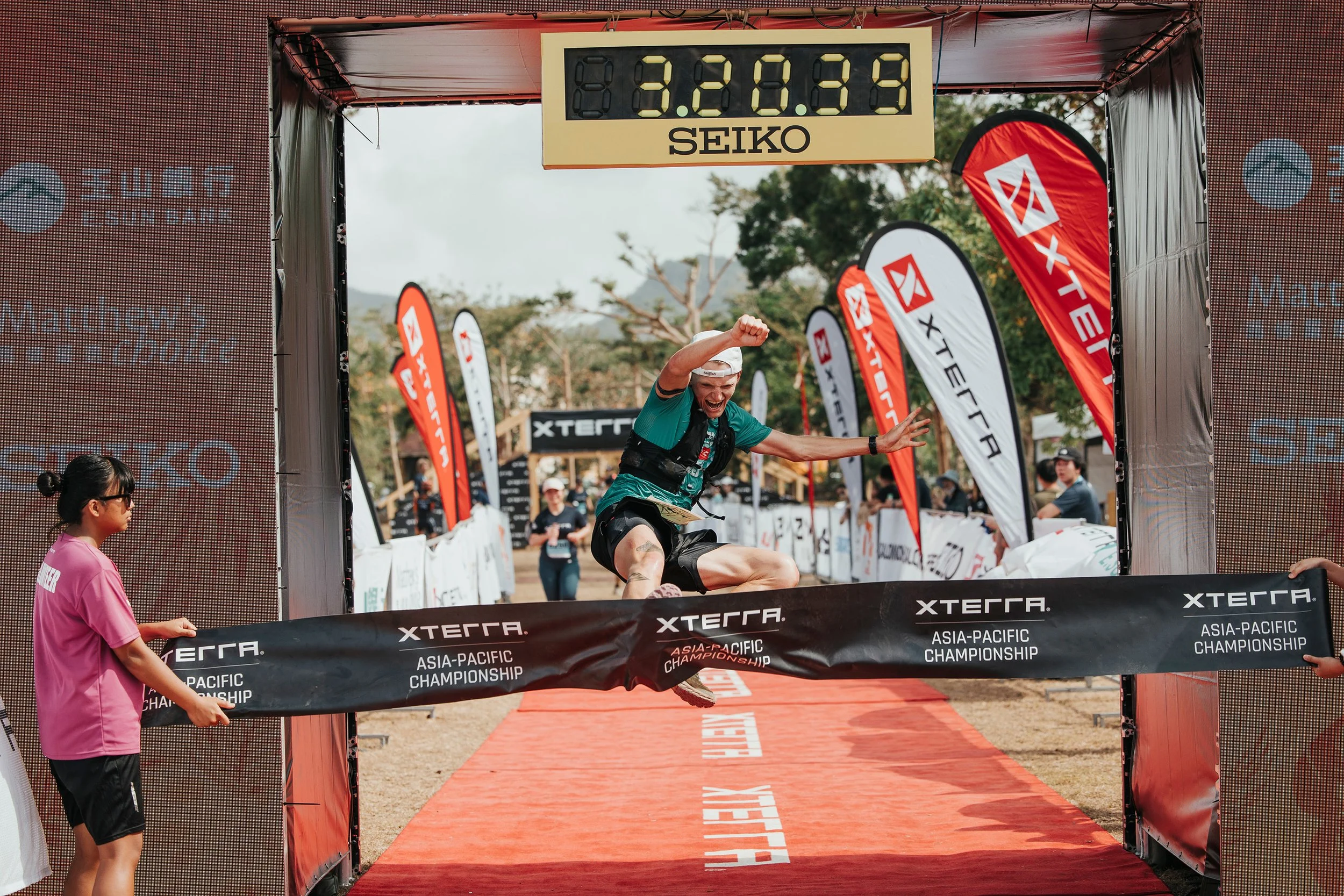 Un homme franchissant la ligne d'arrivée lors d'une compétition d'XTERRA, en pleine course en plein air, sous un ciel nuageux, avec des drapeaux et d'autres participants en arrière-plan.