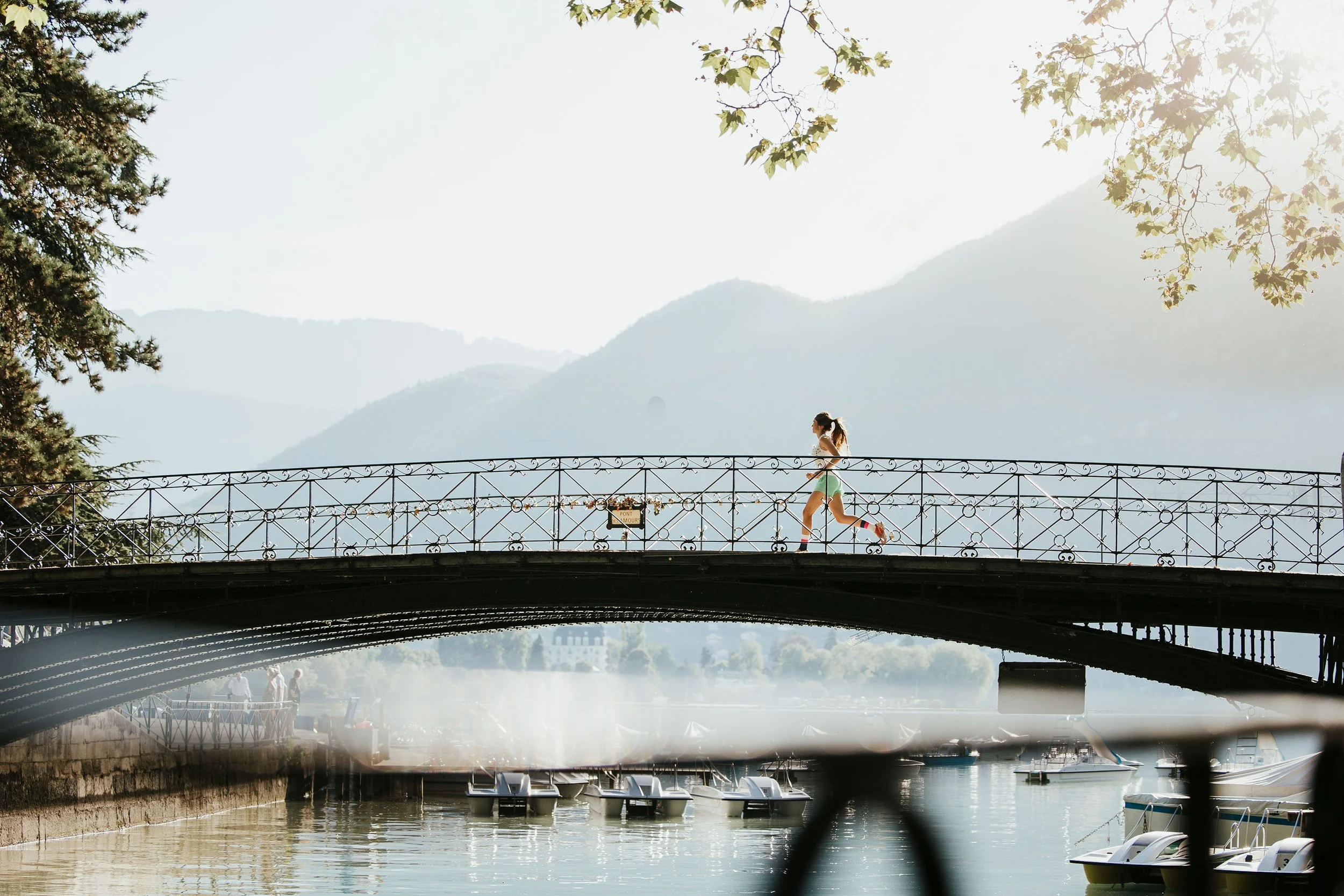 Femme courant sur un pont en métal au-dessus d'un lac avec des voiliers, montagnes en arrière-plan, arbres en premier plan, soleil brillant.