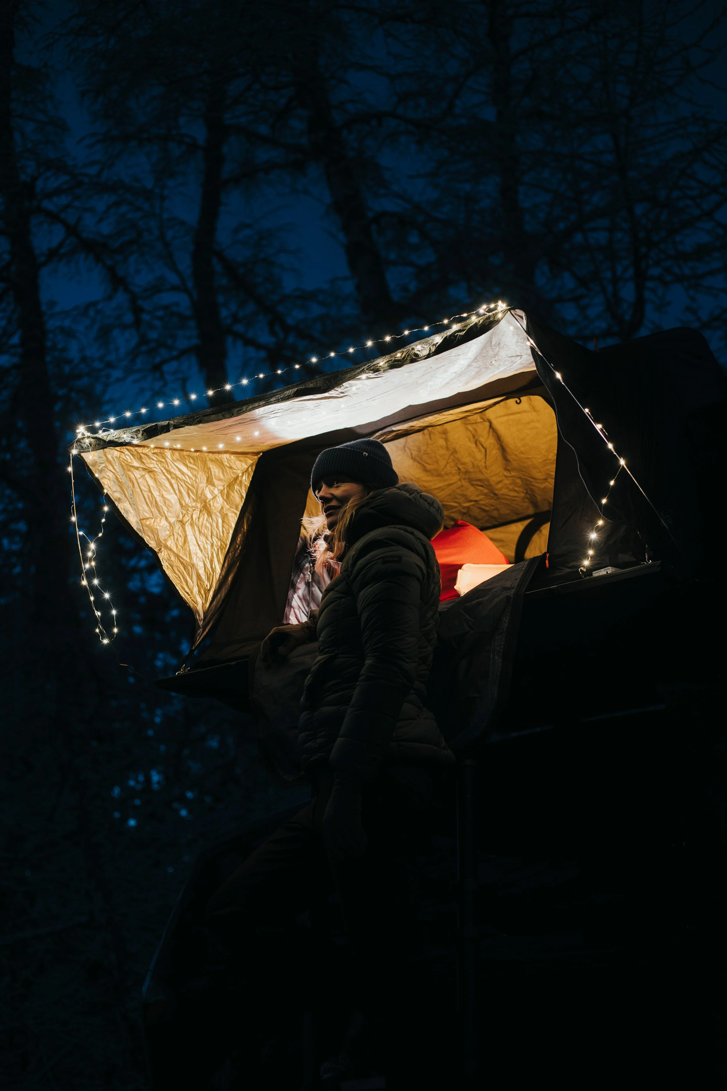 Une femme habillée chaudement dans une cabane suspendue, éclairée par des guirlandes lumineuses, dans une forêt nocturne.