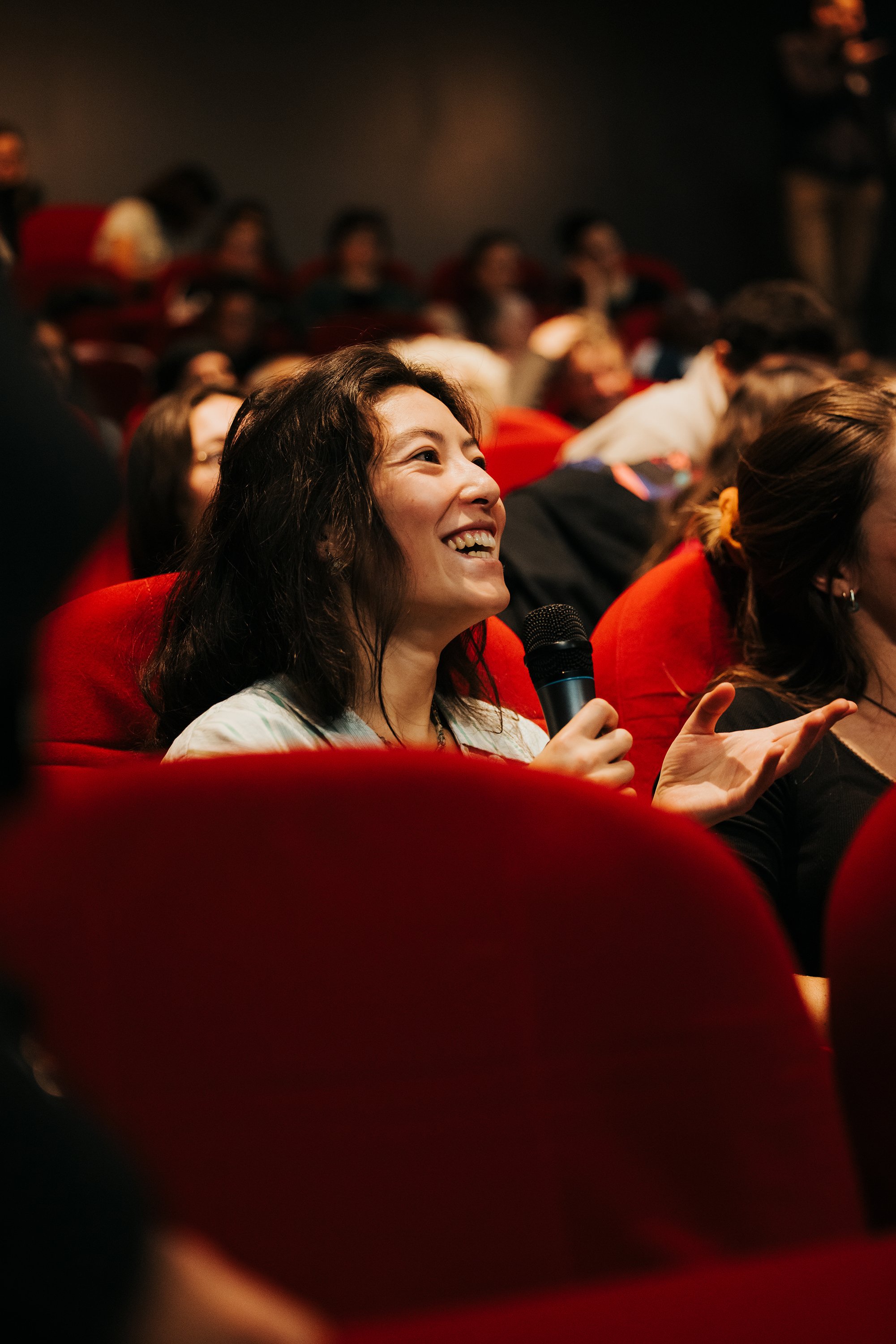 Une femme souriante tenant un microphone lors d'une conférence ou d'un événement, assise dans une salle remplie de personnes.