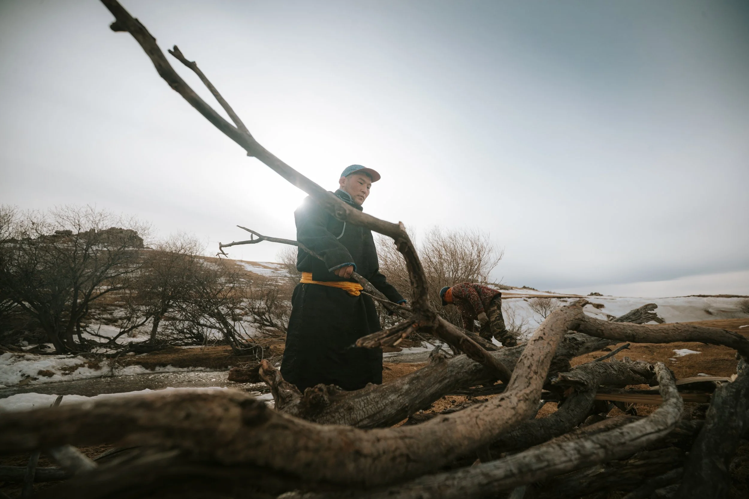 Deux hommes récoltent du bois dans un paysage enneigé avec des arbres dénudés, sous un ciel nuageux