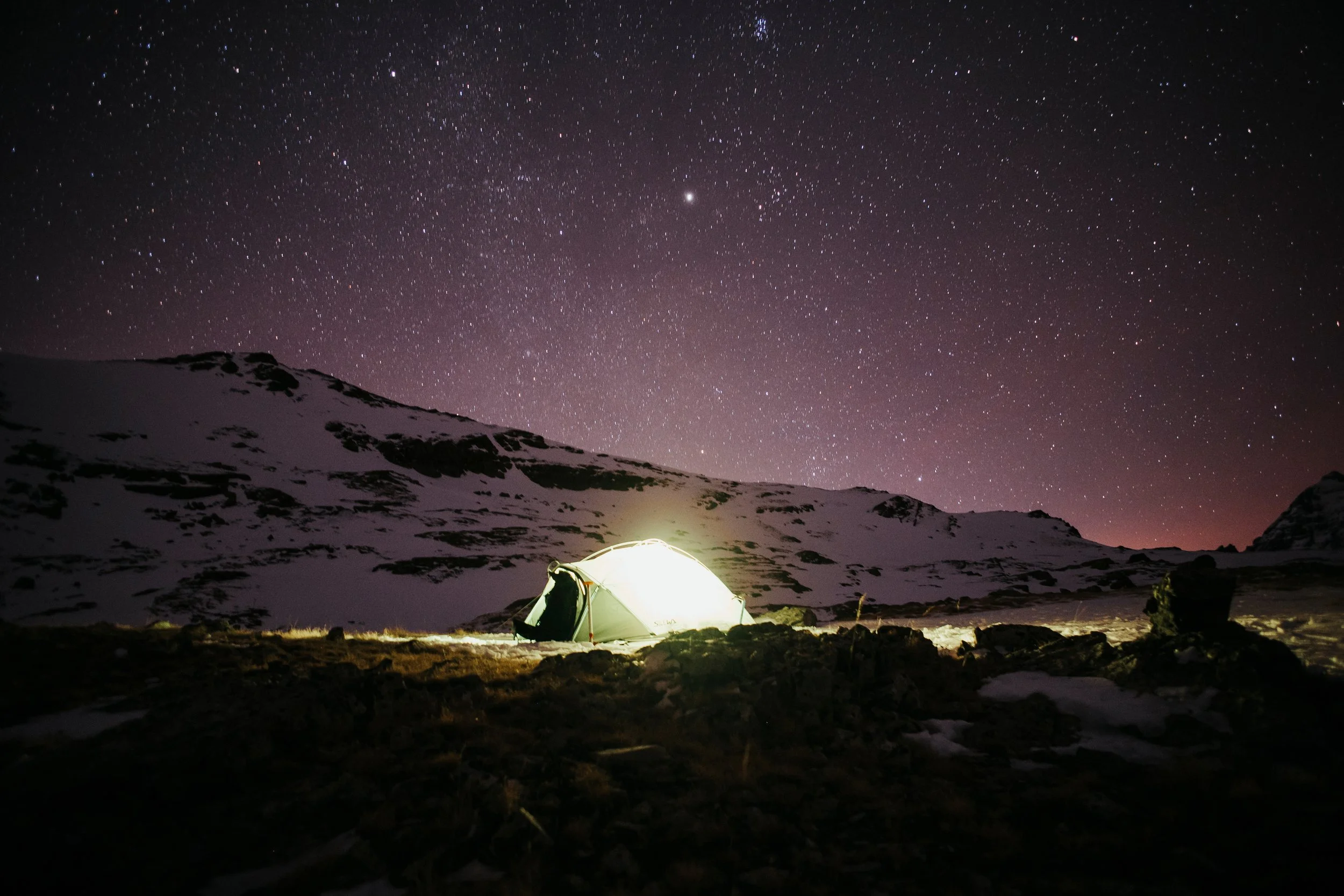 Une tente éclairée sous un ciel étoilé dans un paysage montagneux enneigé.