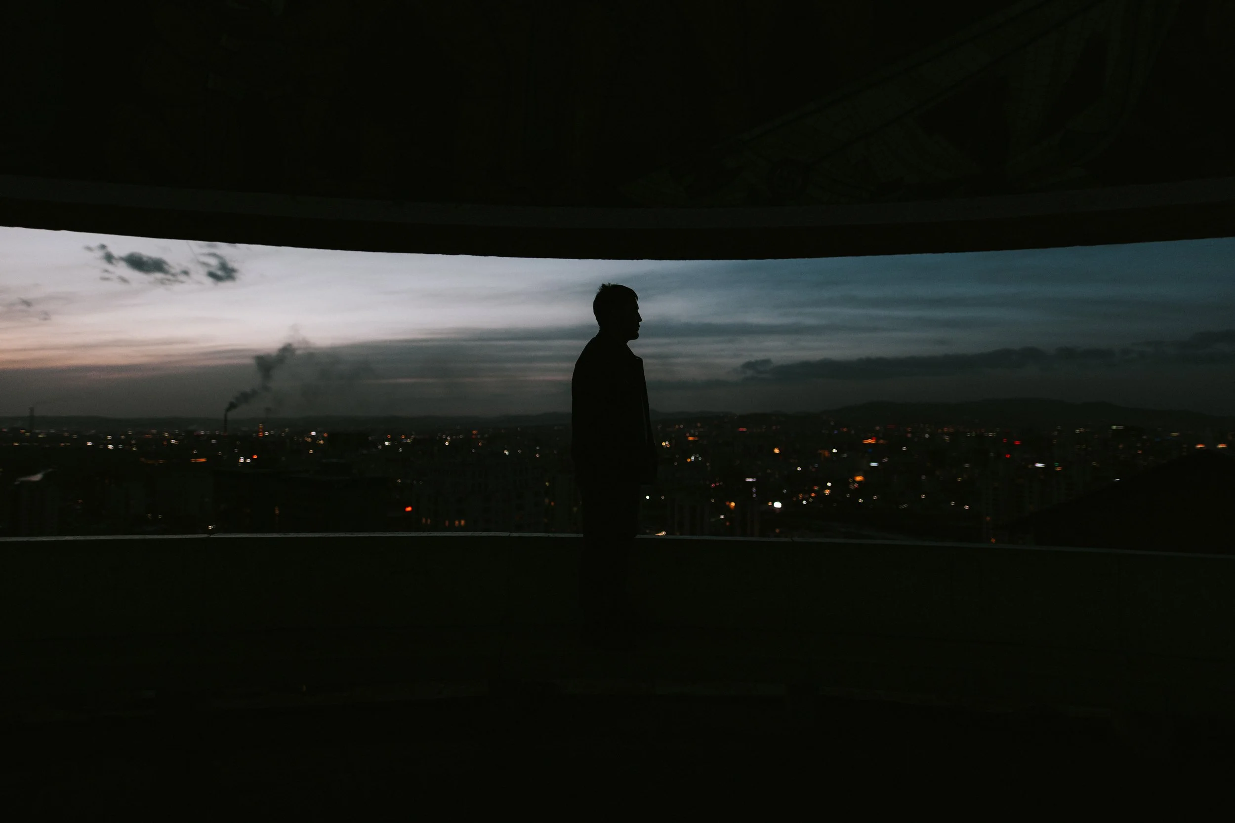 Silhouette d'un homme debout devant une grande fenêtre donnant sur une ville illuminée au crépuscule, avec un ciel nuageux.