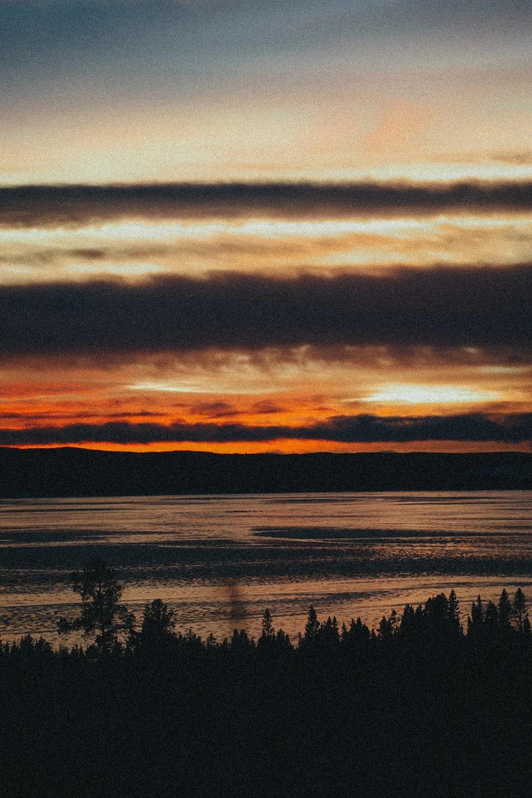 Coucher de soleil avec des nuages colorés au-dessus d’un lac, avec silhouette d’arbres au premier plan.