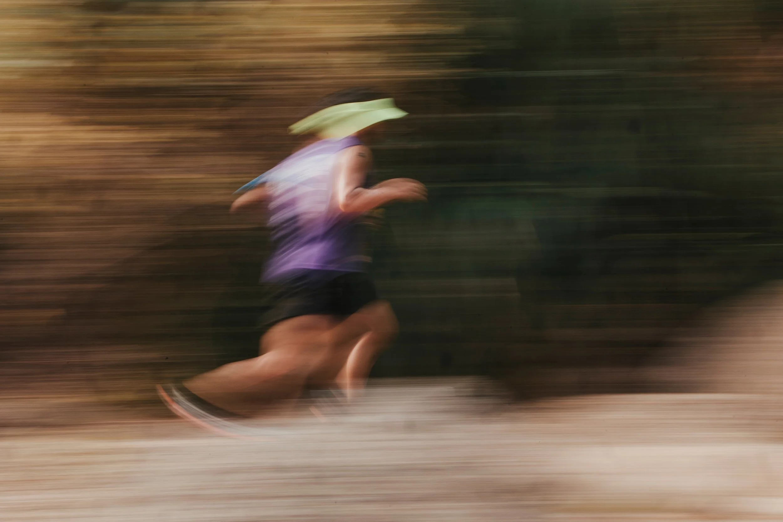 Une personne en course portant un chapeau, vêtue d'un haut violet et d'un short noir, en mouvement rapide sur un chemin de terre, avec un arrière-plan flou de végétation.