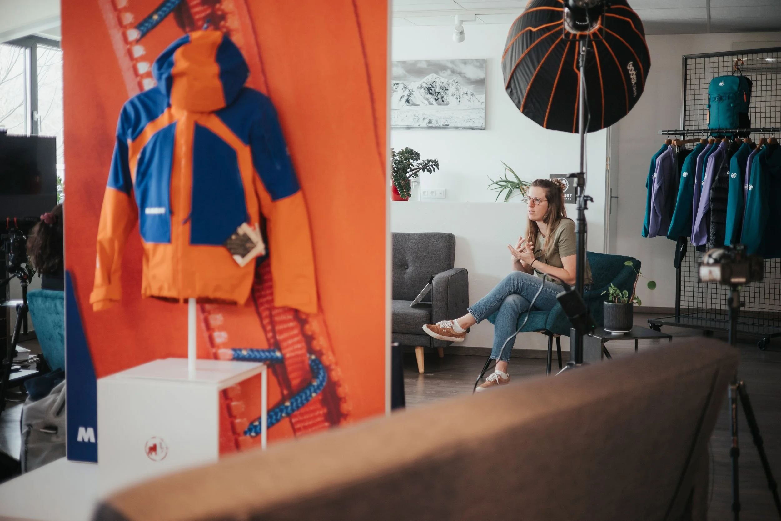 Une personne assise dans une salle d'interview ou de tournage, avec un fond de vêtements et d'équipements MAMMUT, une lumière de studio, et un logo de marque de vêtements sur une veste orange et bleue.