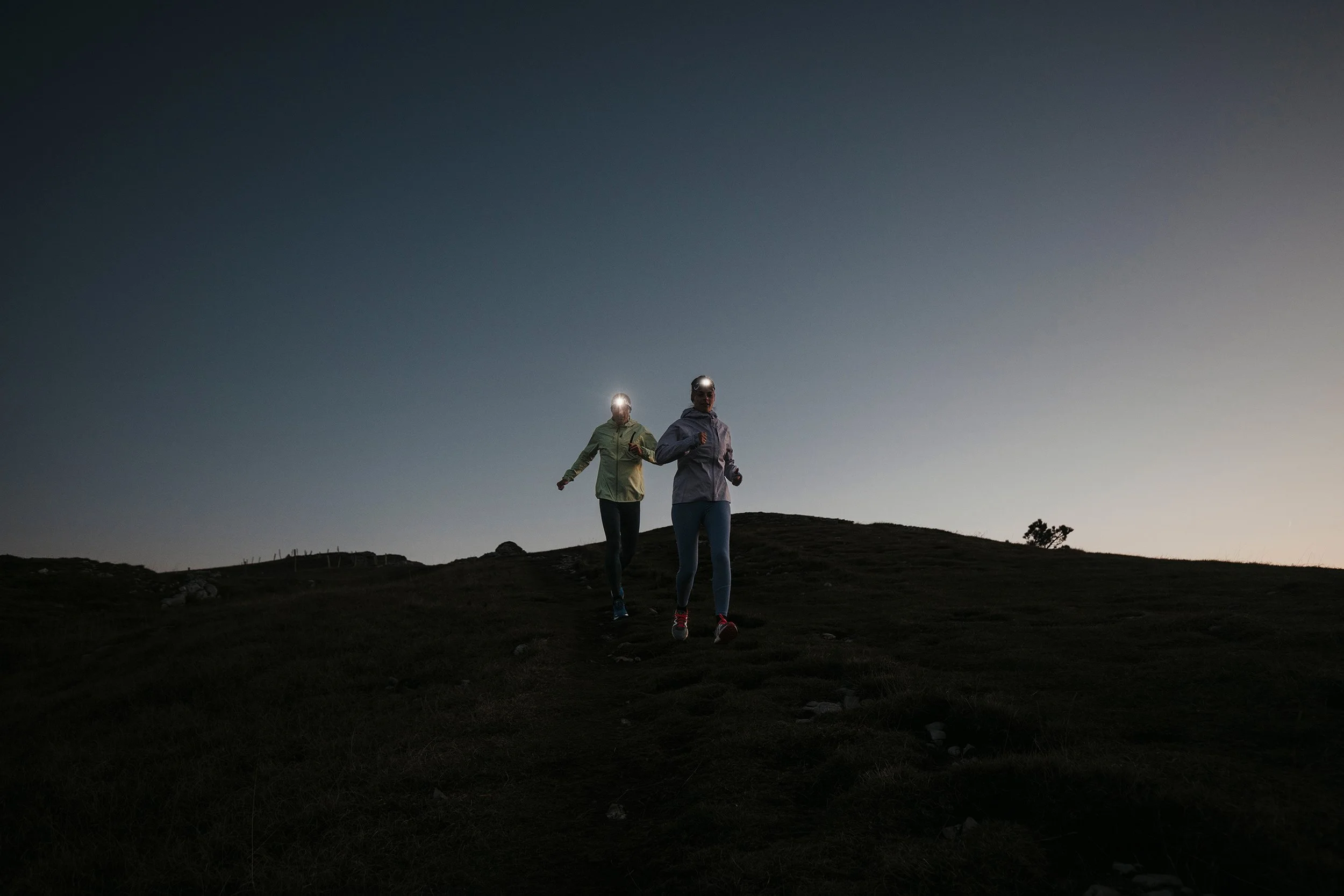 Deux personnes en tenue de sport courant sur une colline au crépuscule, portant des lampes frontales.