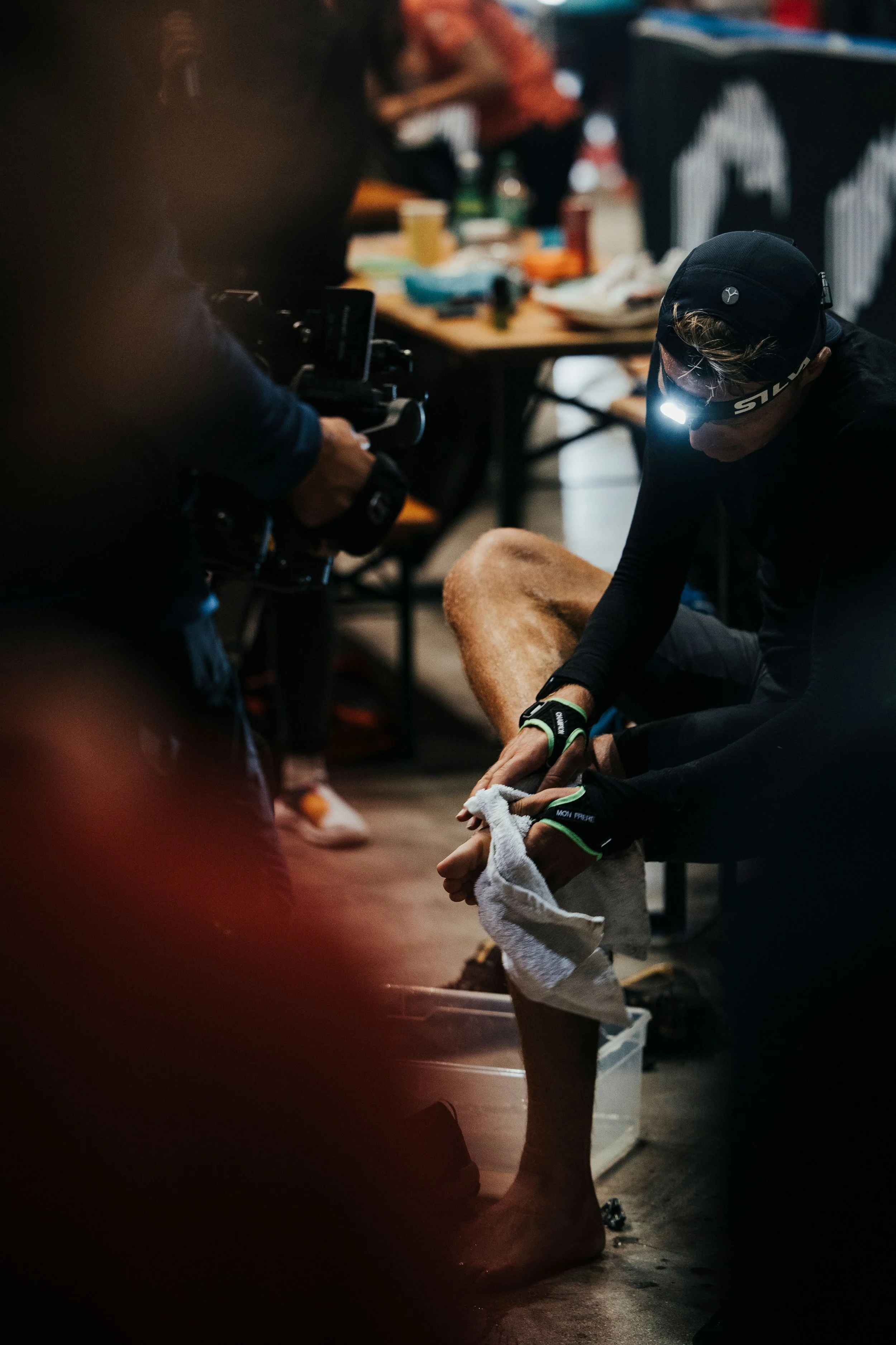 Une personne en tenue de sport, portant une lampe frontale, s'essuyant la jambe avec une serviette dans une pièce sombres, avec une table en arrière-plan remplie d'objets divers.