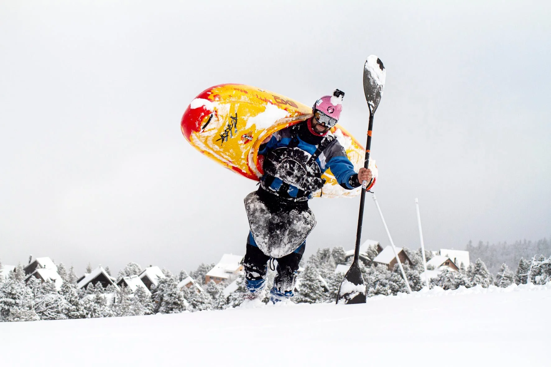 Eric Deguil en tenue de kayak, avec casque et pagaie, marchant dans la neige en terrain montagneux enneigé.
