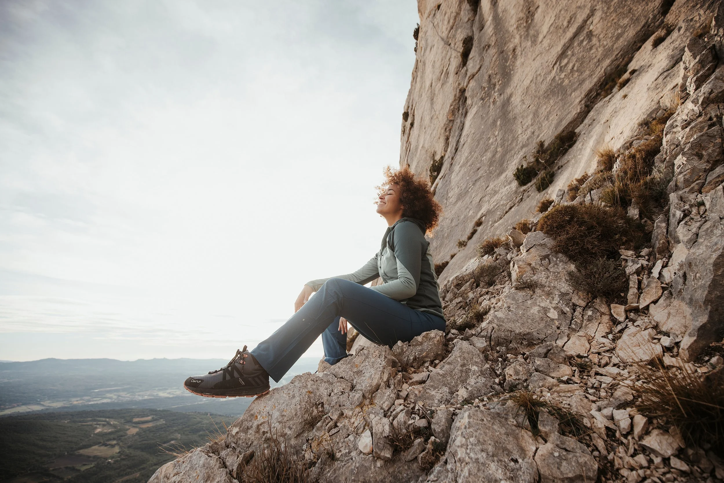 Femme souriante en tenue de randonnée assise sur un rocher en montagne, face au soleil couchant, avec vue sur la vallée en contrebas.