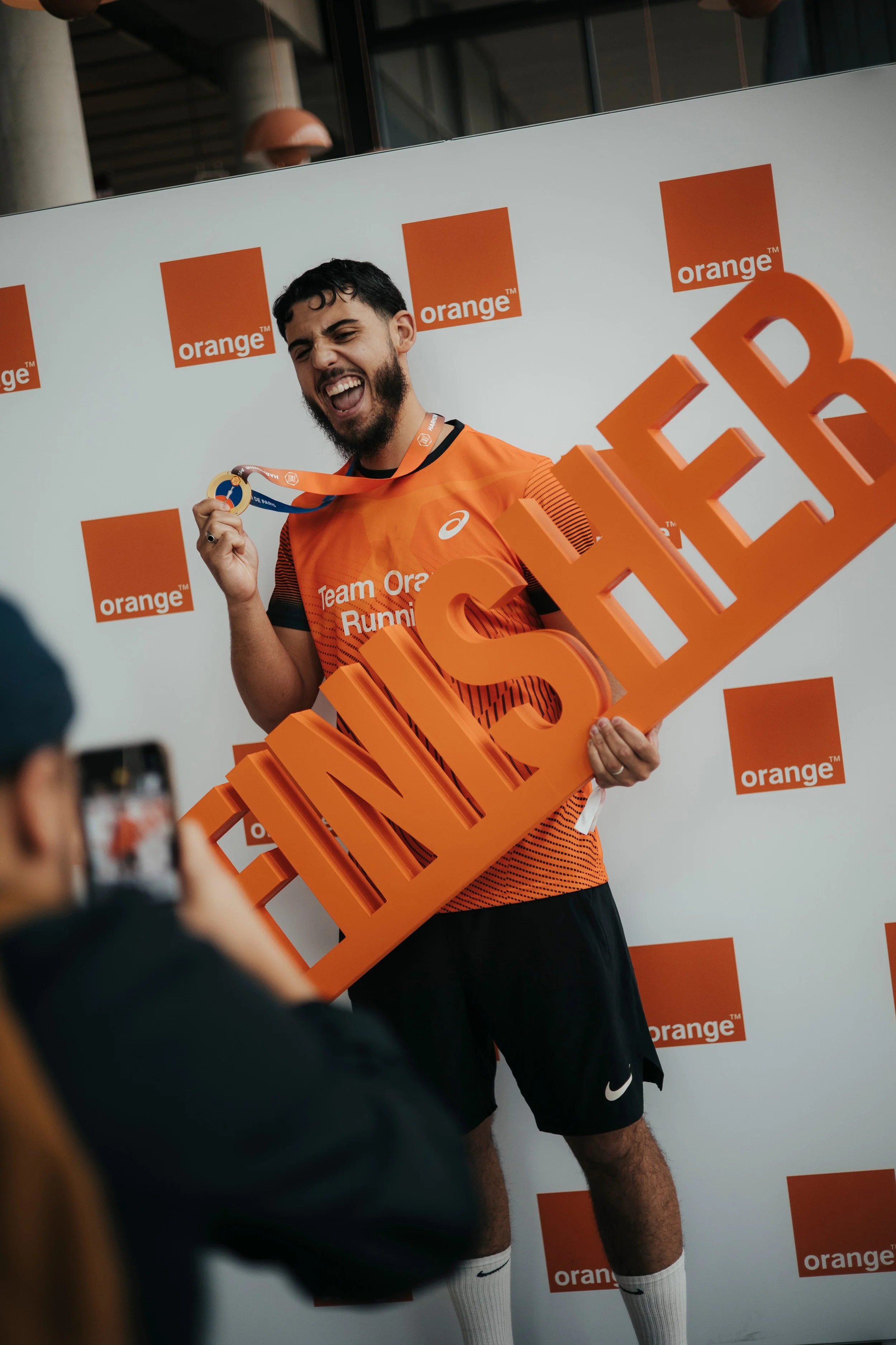 Un homme souriant portant une médaille et tenant un grand panneau orange avec le mot « FINISHER » lors d'une cérémonie de remise de médaille, avec un fond blanc decoré de logos « orange ». Il porte un t-shirt orange avec l'inscription « Team Orange R