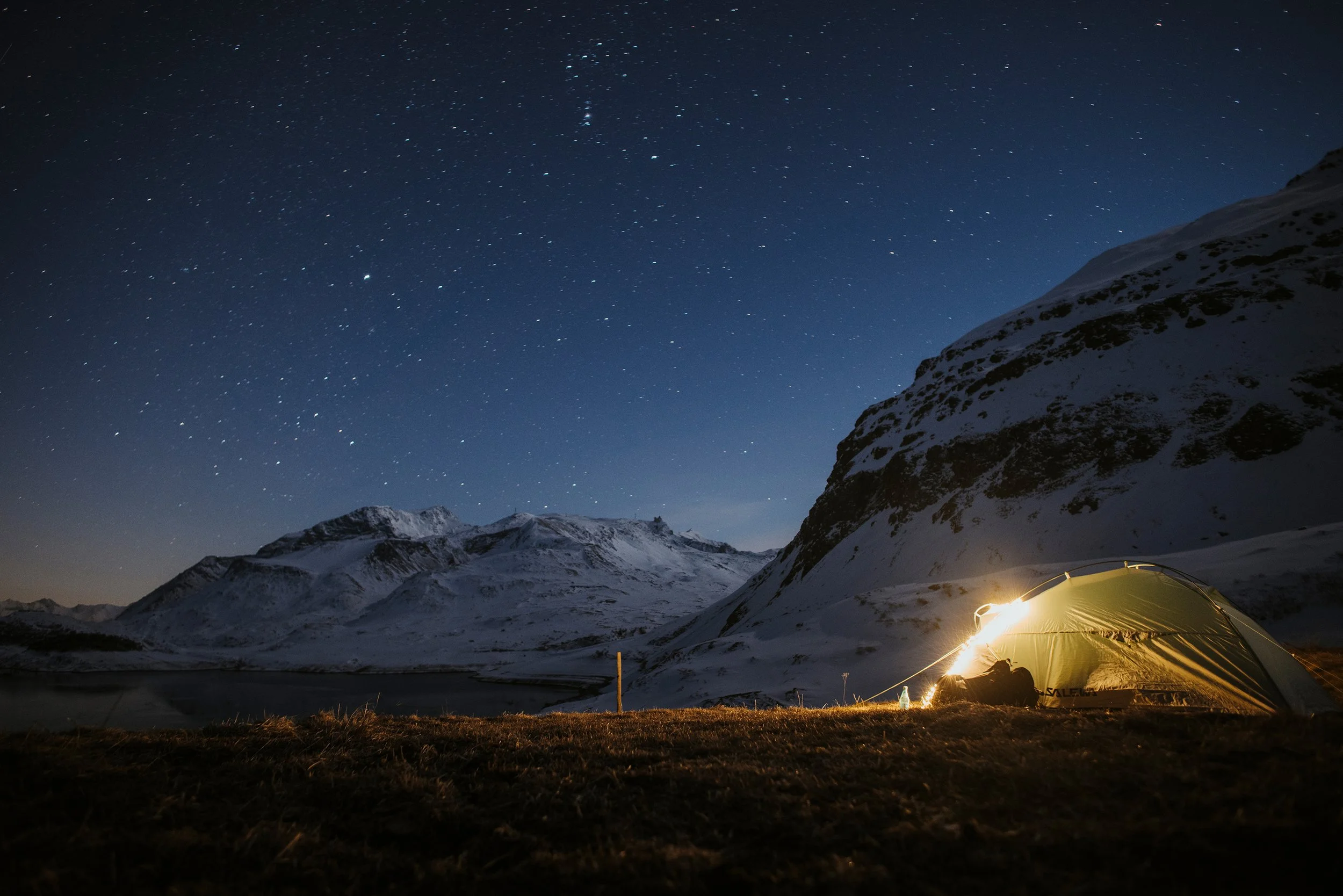 Tente éclairée dans un paysage montagneux enneigé sous un ciel étoilé la nuit.