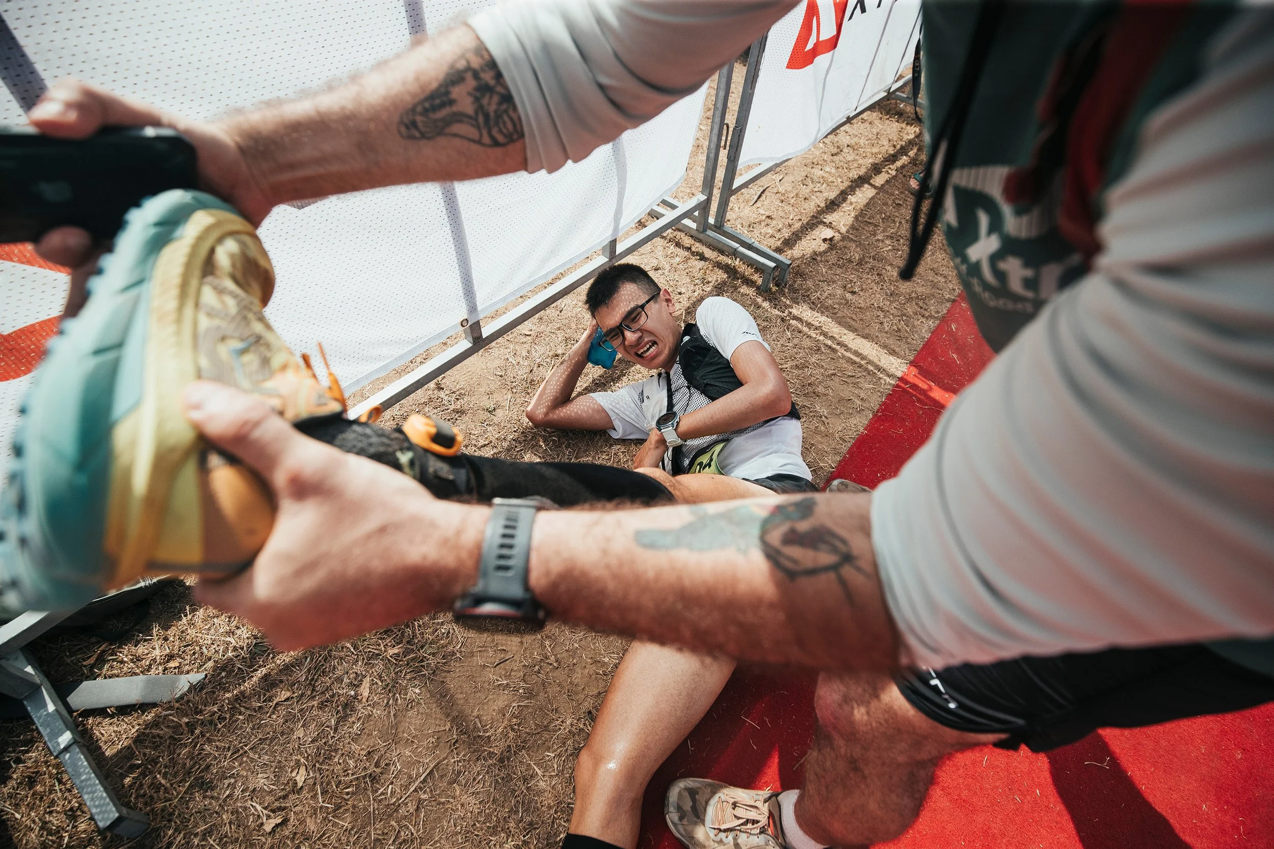 Un jeune homme semble avoir glissé et est assis par terre, tenant sa tête avec une expression de douleur lors d'une course, avec d'autres personnes autour. L'environnement semble être une piste en terre.