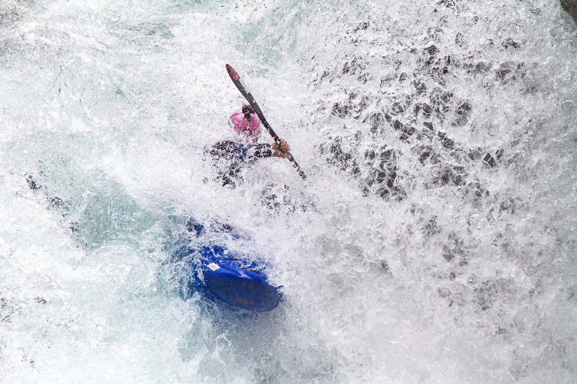 Une personne, Eric Deguil, en kayak équipée d'un casque rose navigue dans une rivière agitée avec beaucoup d'écume et de vagues.