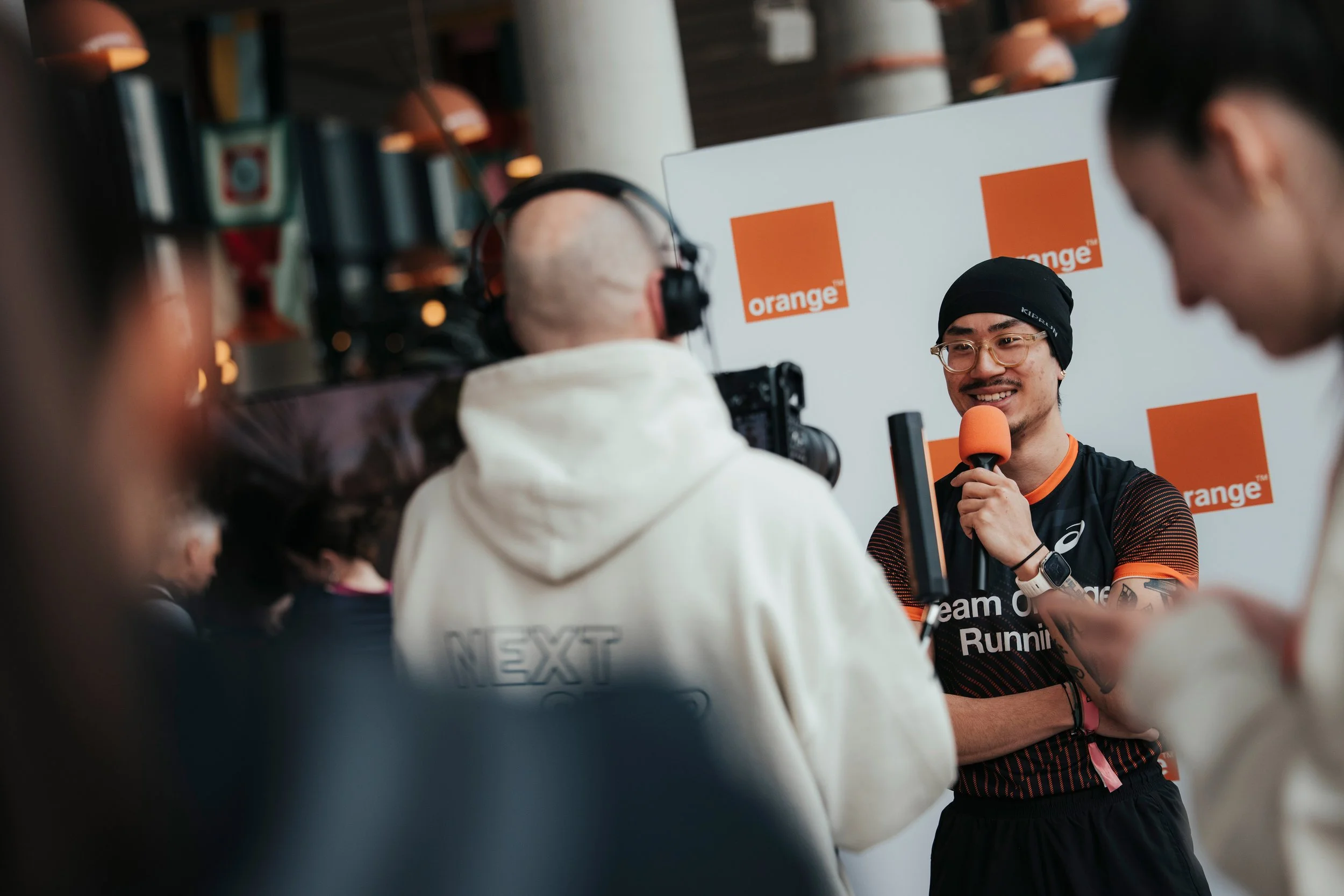 Un homme souriant lors d'une interview, tenant un micro orange avec un fond arborant le logo d'Orange, porteur d'une tenue de course et portant un bonnet noir.