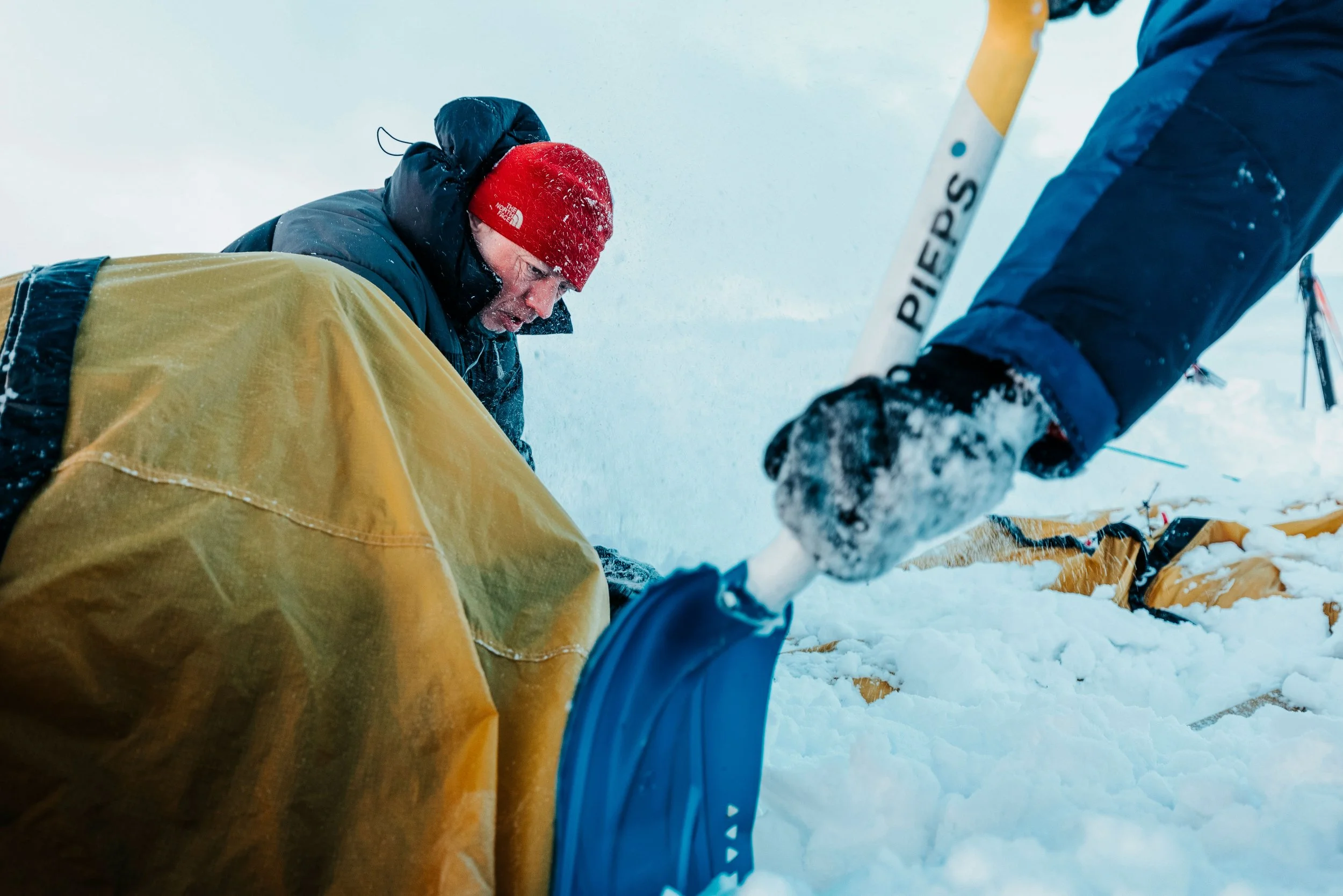 Une personne portant un bonnet rouge et une veste noire aide une autre personne, dont la main est visible, à sortir de la neige par temps froid, avec un équipement de ski visible à l'arrière.