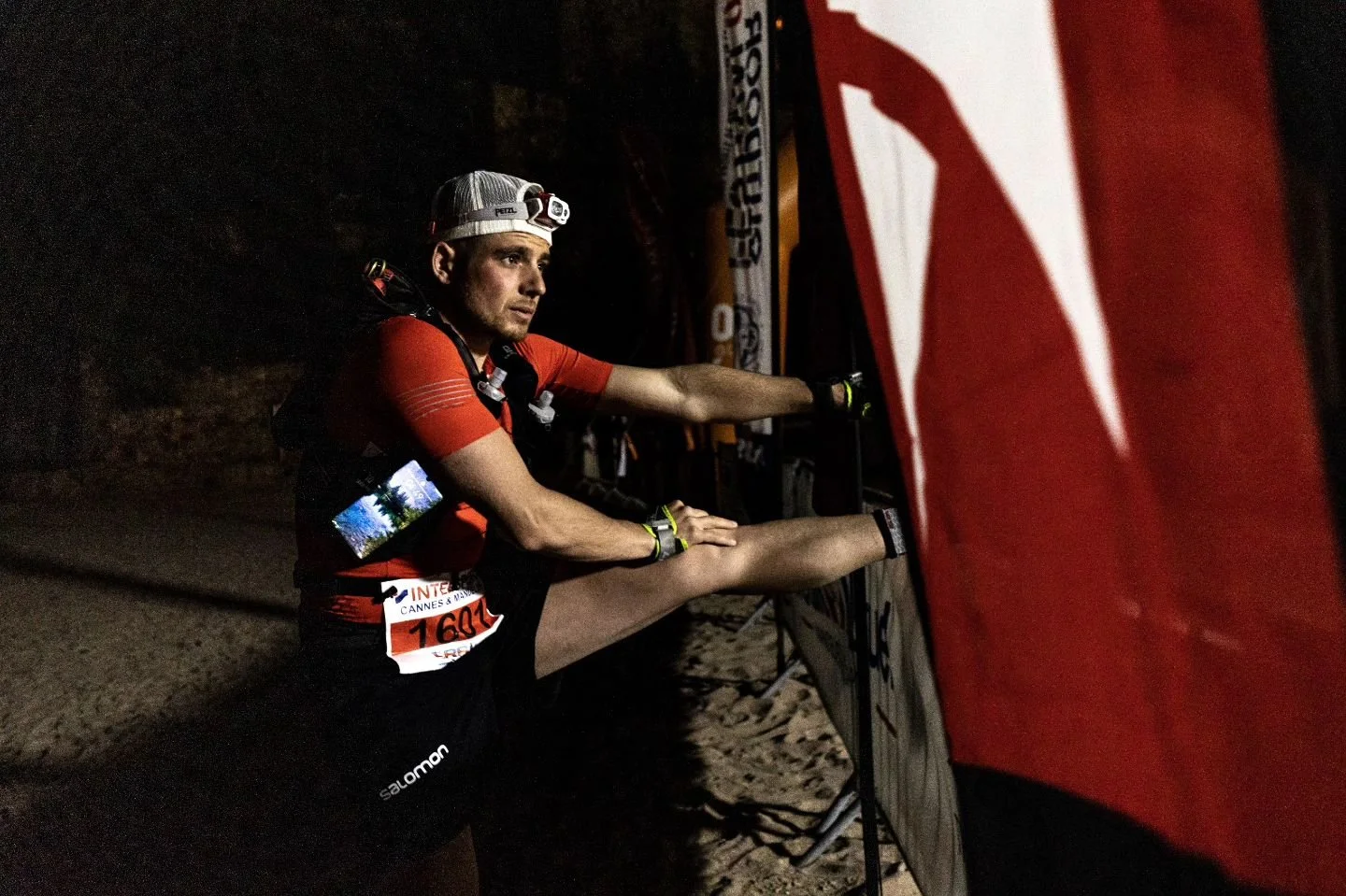 Un homme en tenue de course, portant une montre connectée et une headlamp, s'étire ou se prépare avant une course de nuit sur un terrain non pavé.