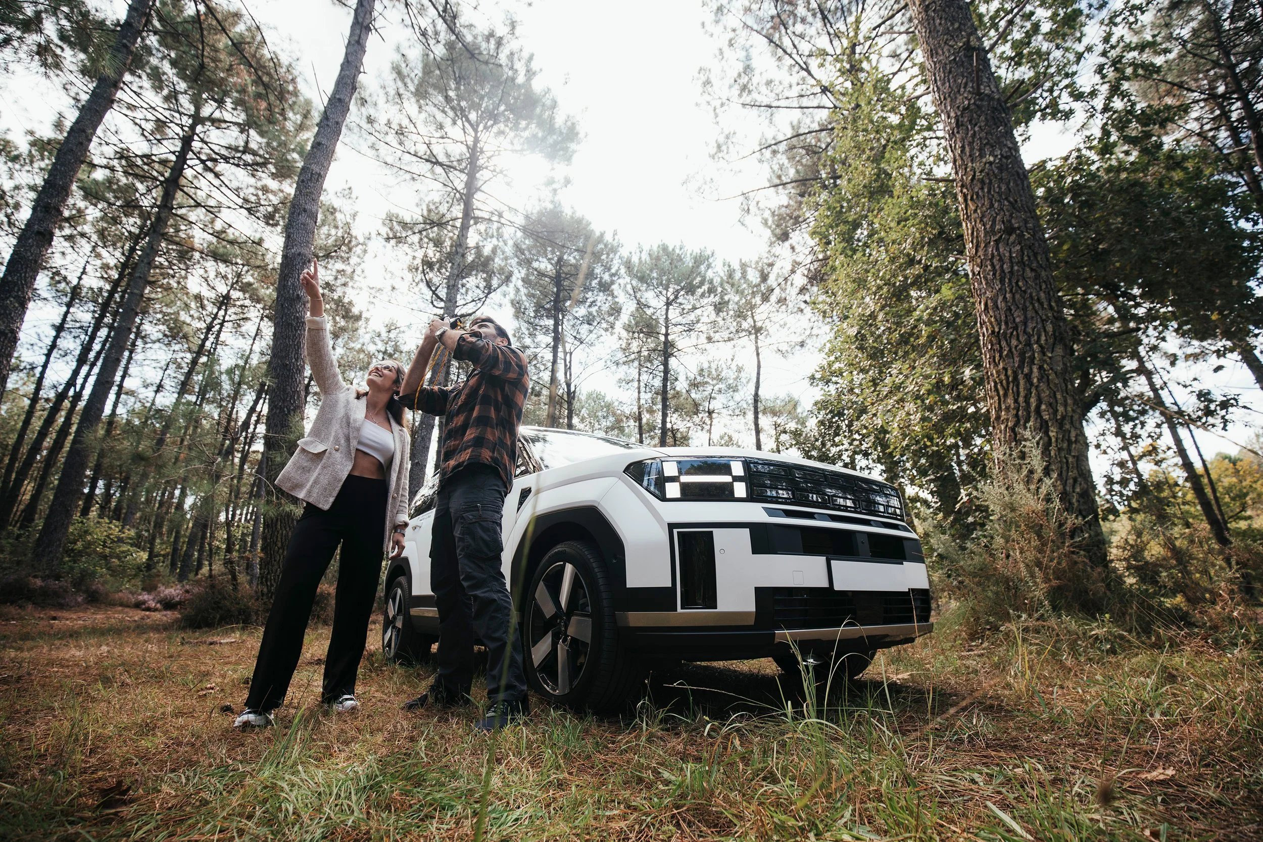 Deux personnes prennent une photo à côté d'un SUV en forêt, avec des arbres et le soleil brillant à l'arrière-plan.