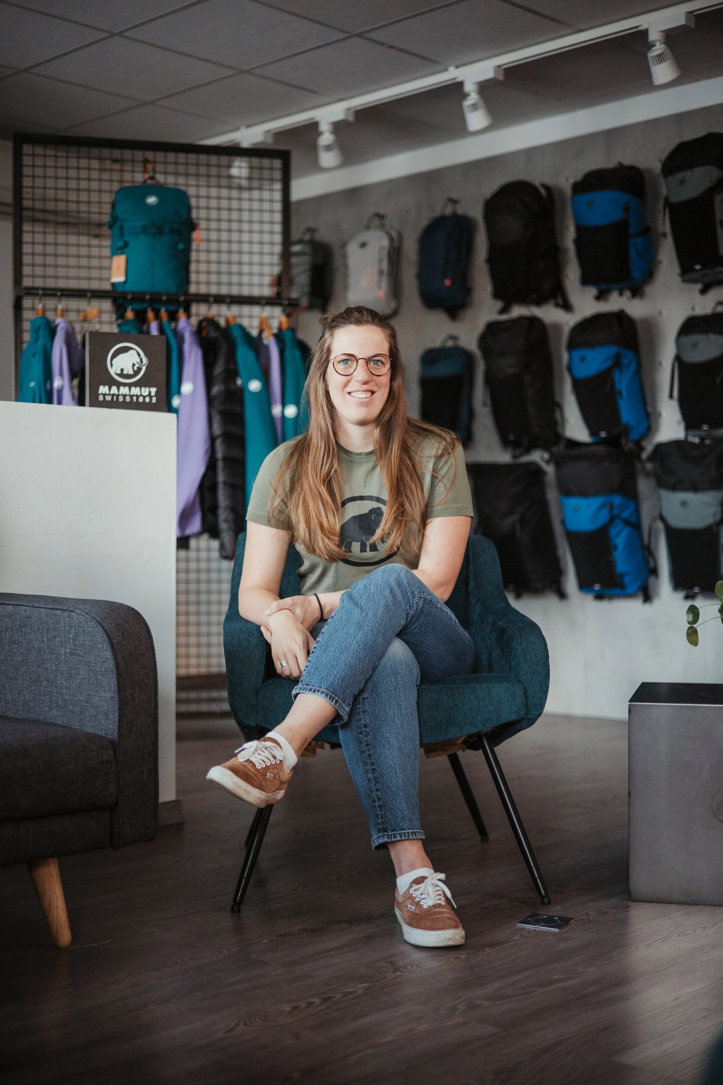 Une femme assise dans un magasin d'équipements de plein air, entourée de sacs à dos et de vêtements de randonnée MAMMUT.