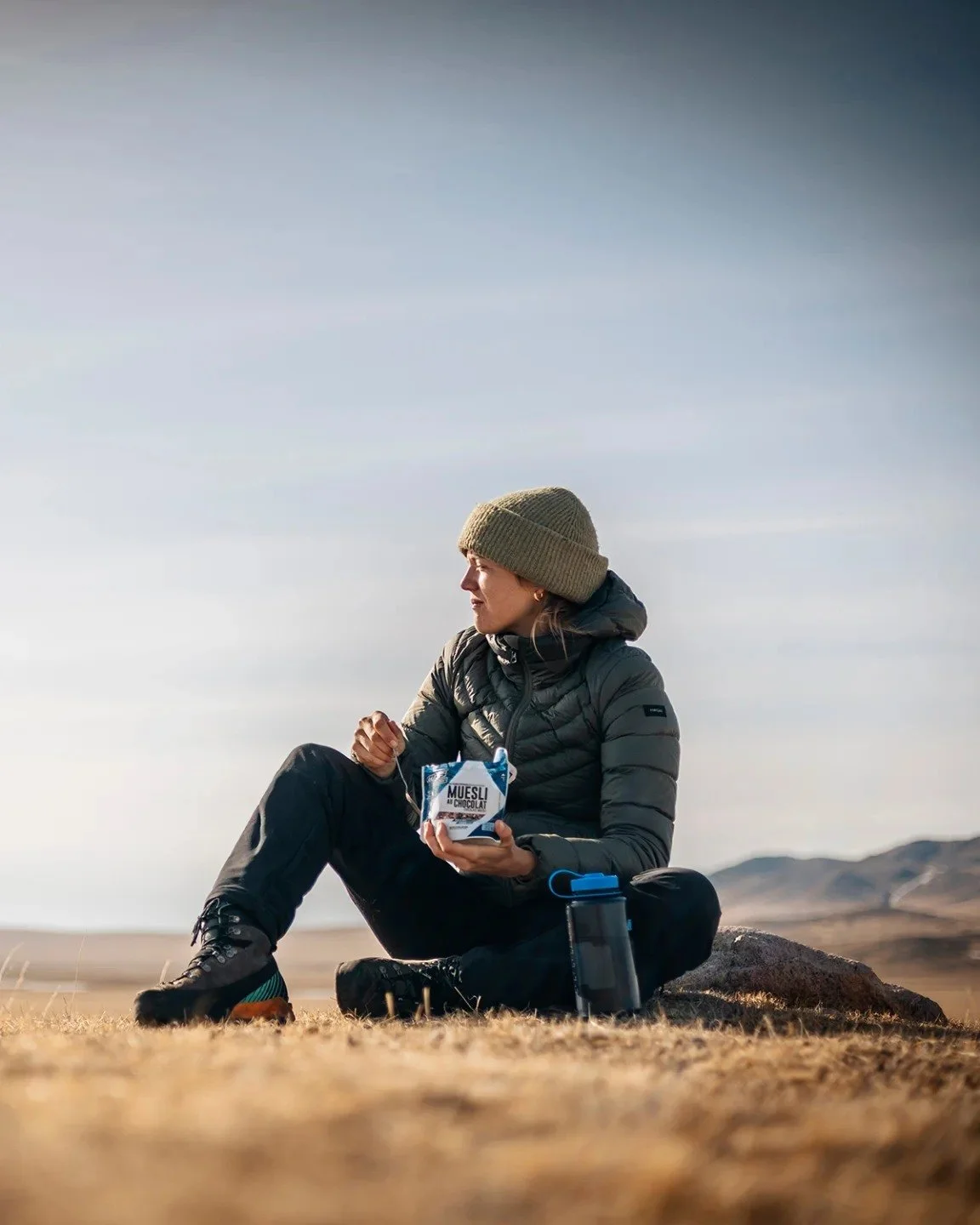 Une femme assise sur le sol dans un environnement désertique avec des montagnes en arrière-plan, mangeant un snack qu'elle tient dans la main, portant un bonnet vert, une veste grise, et une gourde à côté d'elle.
