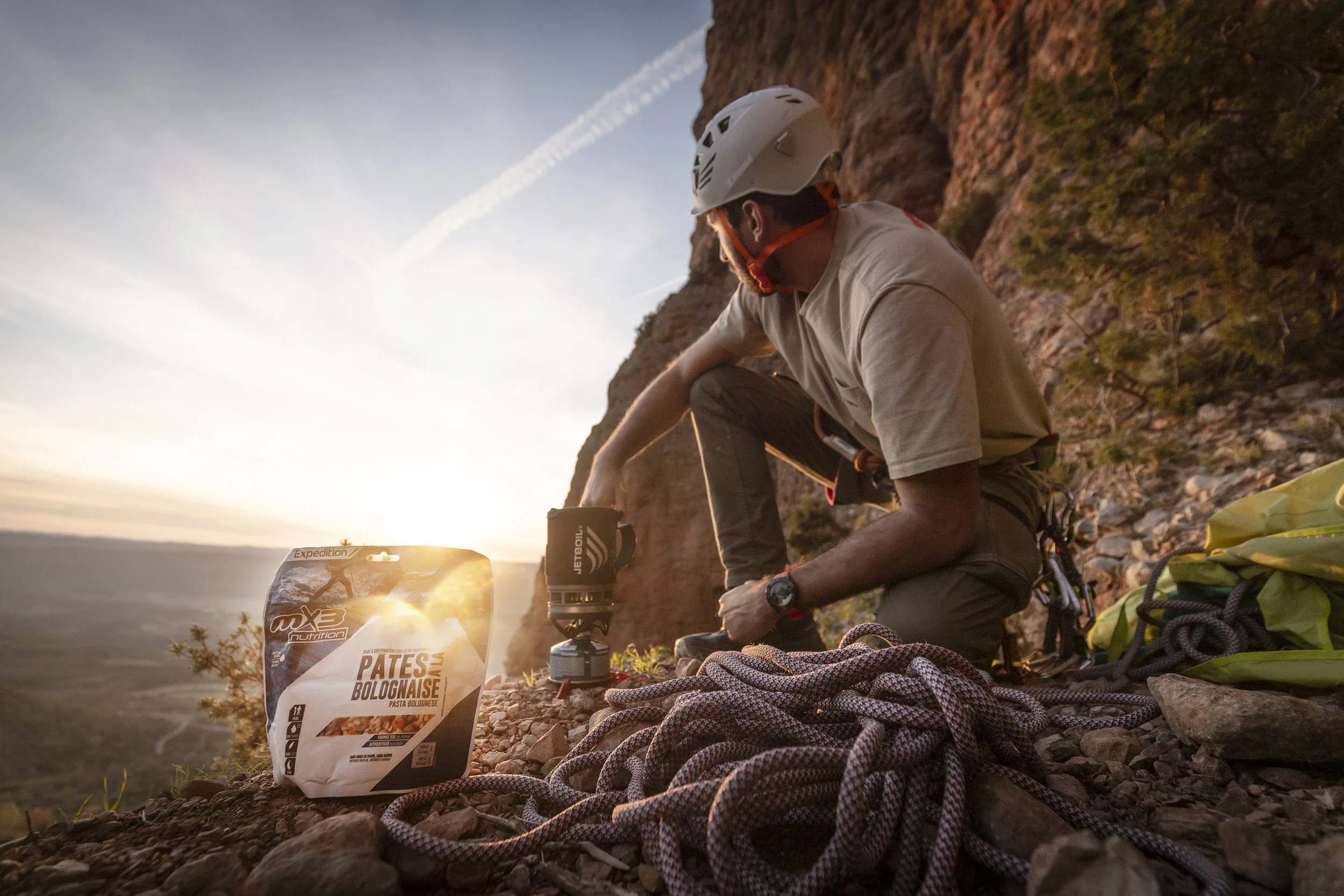 Un homme en short et casque de randonnée préparant un repas avec une lampe de camping sur une falaise rocheuse au coucher du soleil, avec un sac à dos et une corde en premier plan.