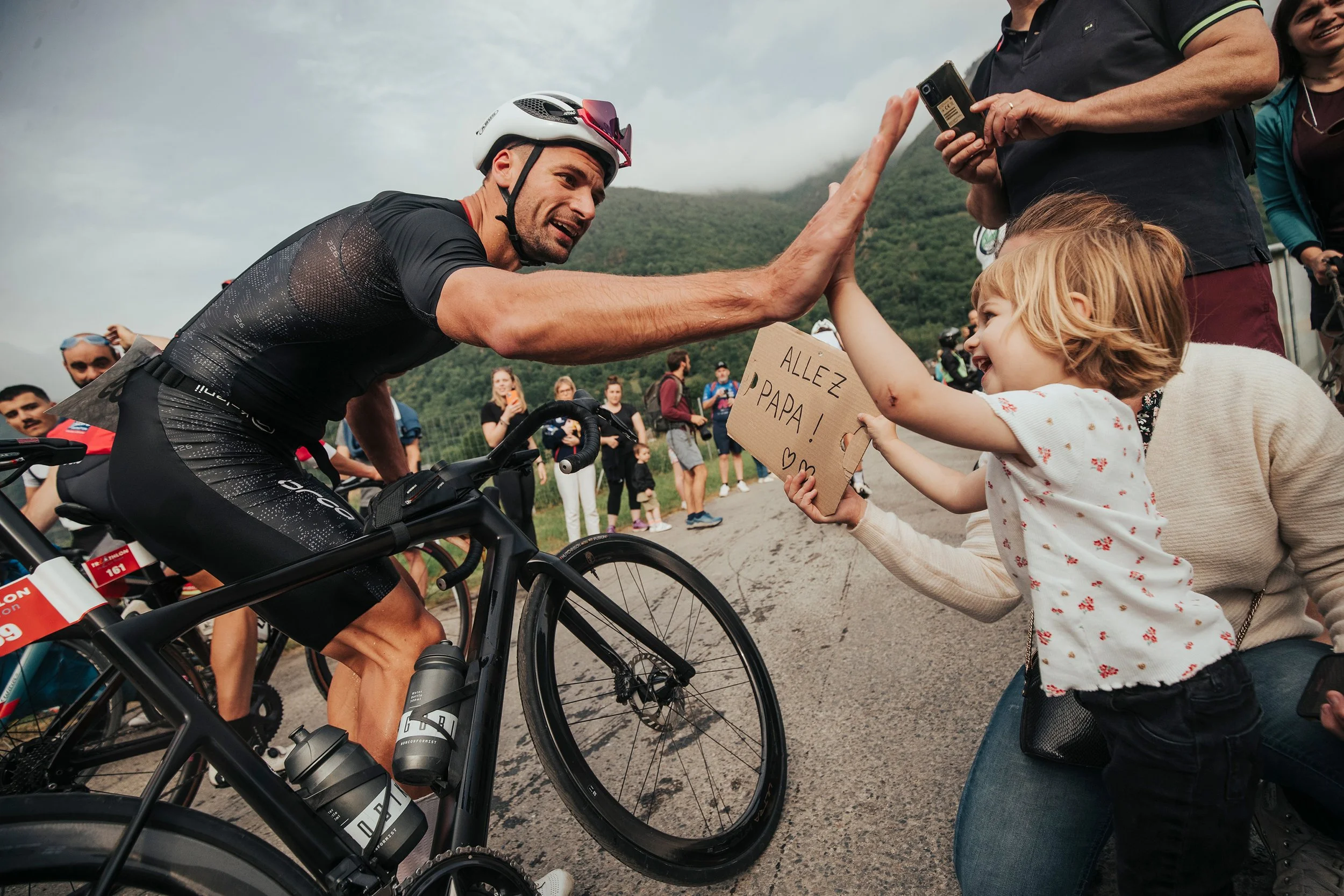 Un homme en tenue de cyclisme échange un high-five avec une jeune fille tenant une pancarte disant "Allez PAPA!" lors d'une course cycliste en plein air, avec un groupe de spectateurs en arrière-plan.