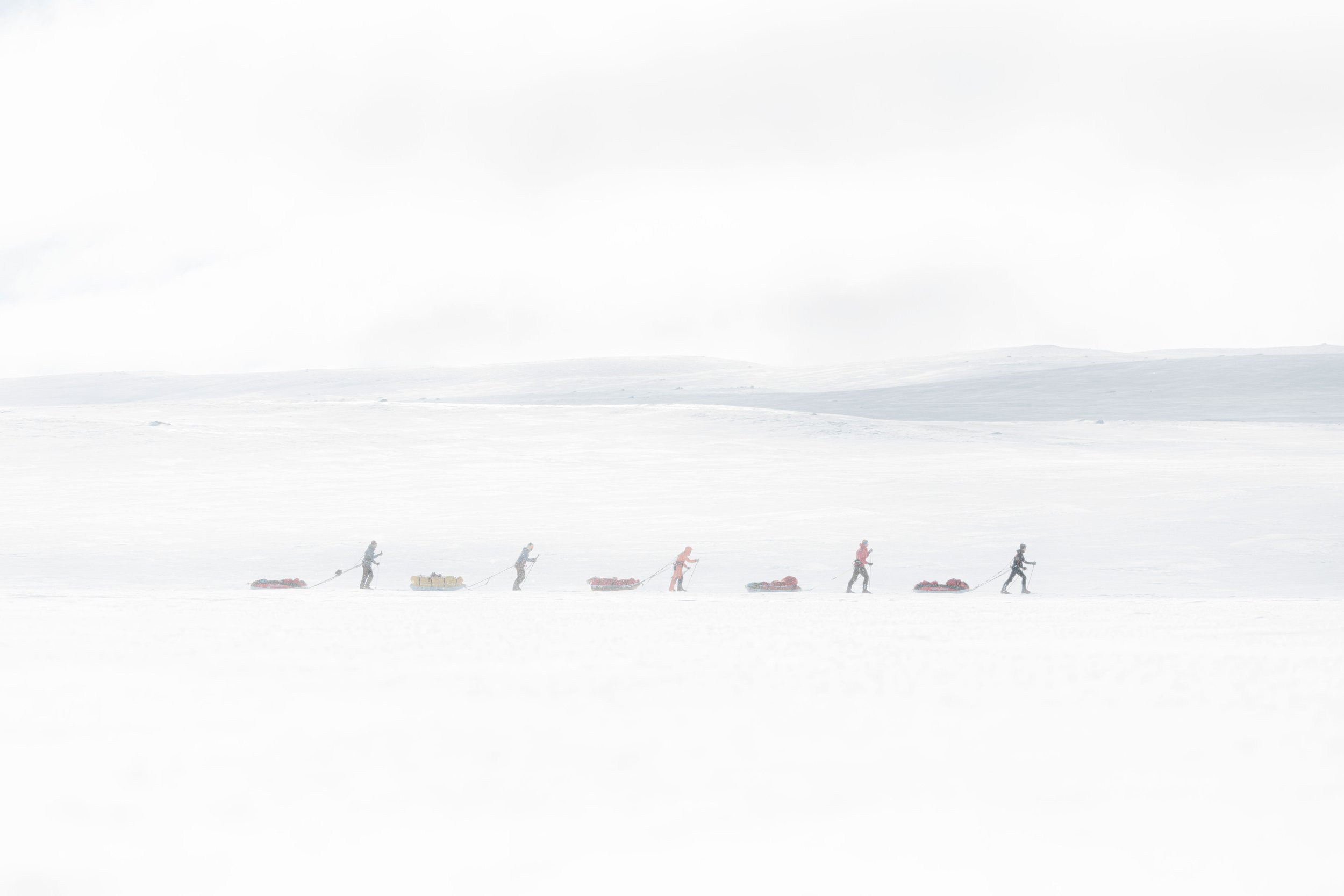 Personnes en skis et pulkas traversant un paysage enneigé en file indienne, tirant des traîneaux chargés de matériel sous un ciel nuageux.